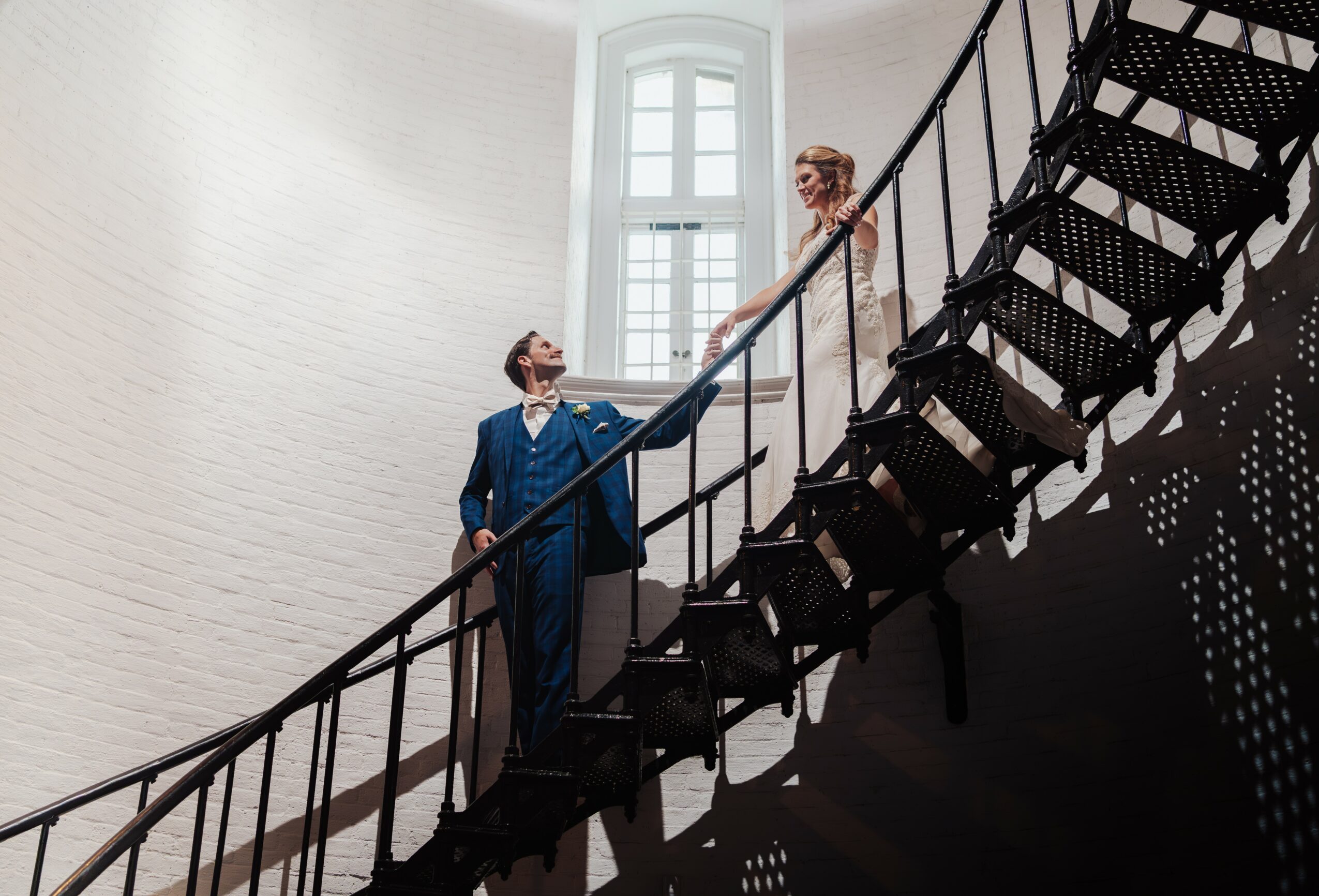Two people, a man and a woman, stand on a staircase inside a building with large windows, sunlight streaming in.