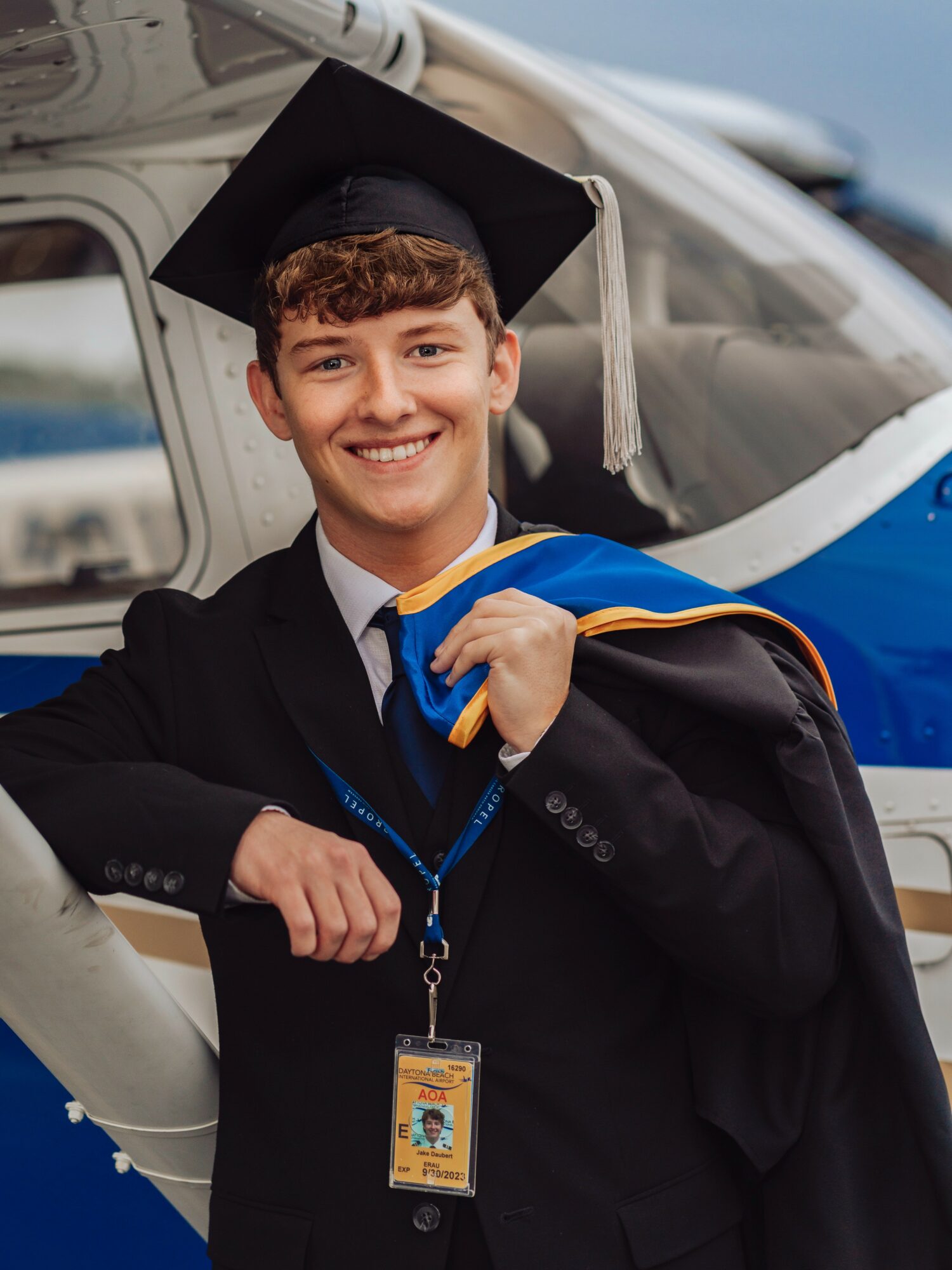Young man in graduation gown and cap smiling, holding a diploma, standing in front of a helicopter.