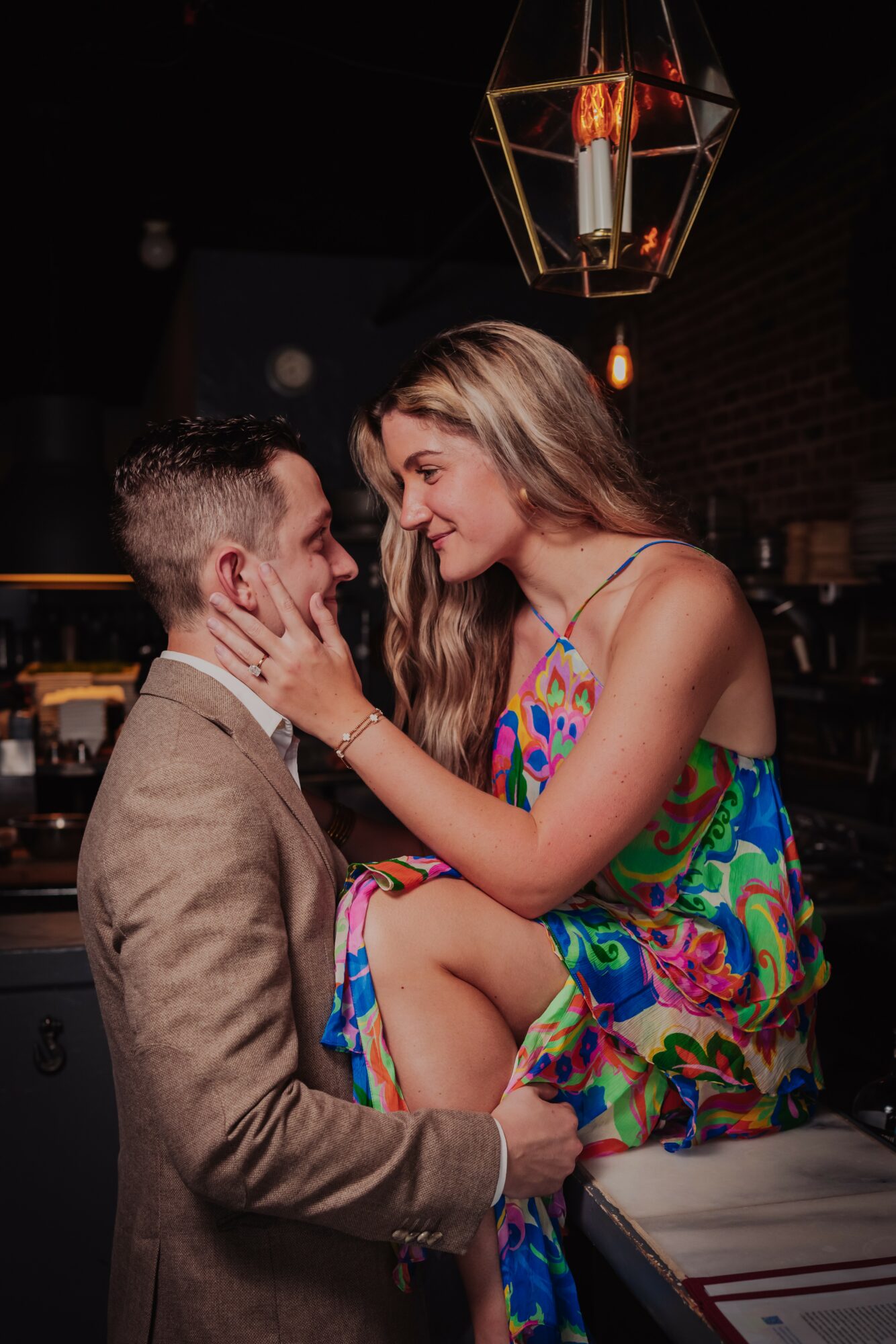 A woman and a man face each other closely, woman touching man's face, in a dimly lit setting with a geometric pendant light above.
