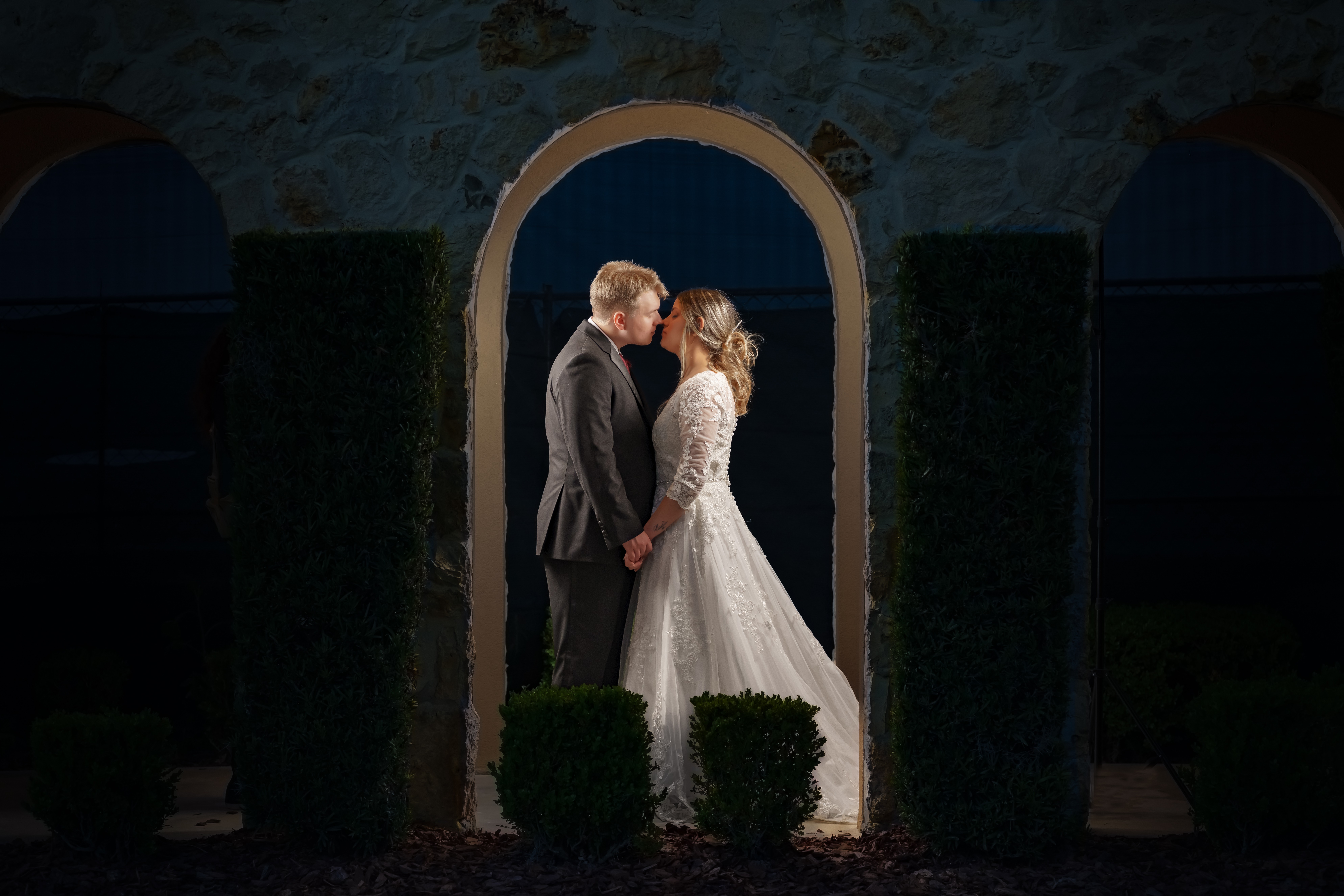 A couple in wedding attire stands close, facing each other, framed by an archway with dark background.