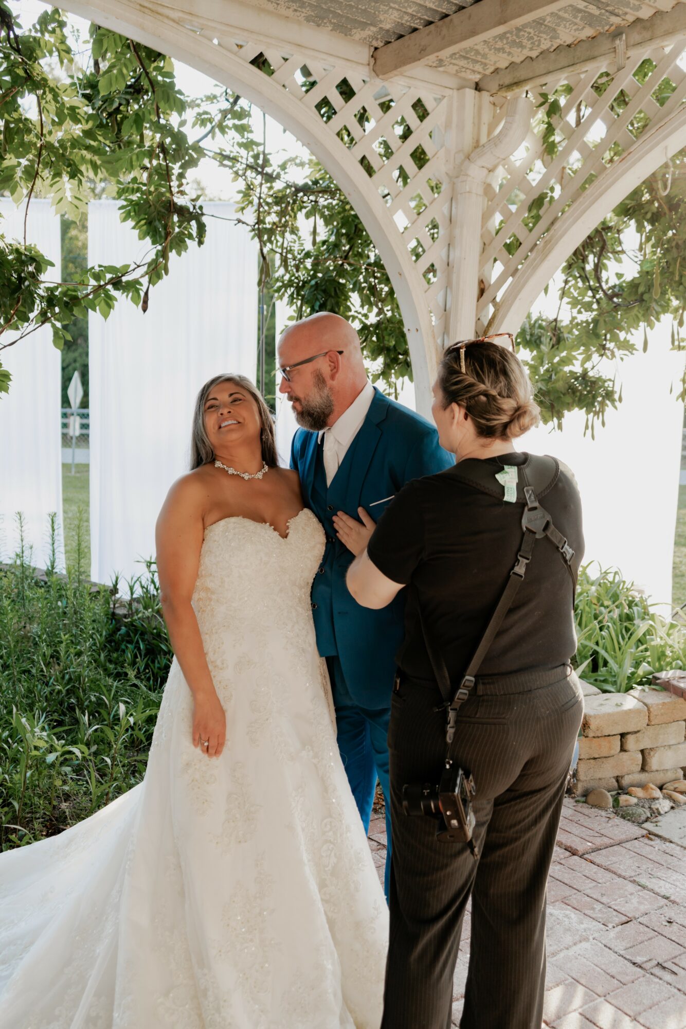 Three people under a white gazebo, one in a wedding dress, smiling and talking, greenery in background.