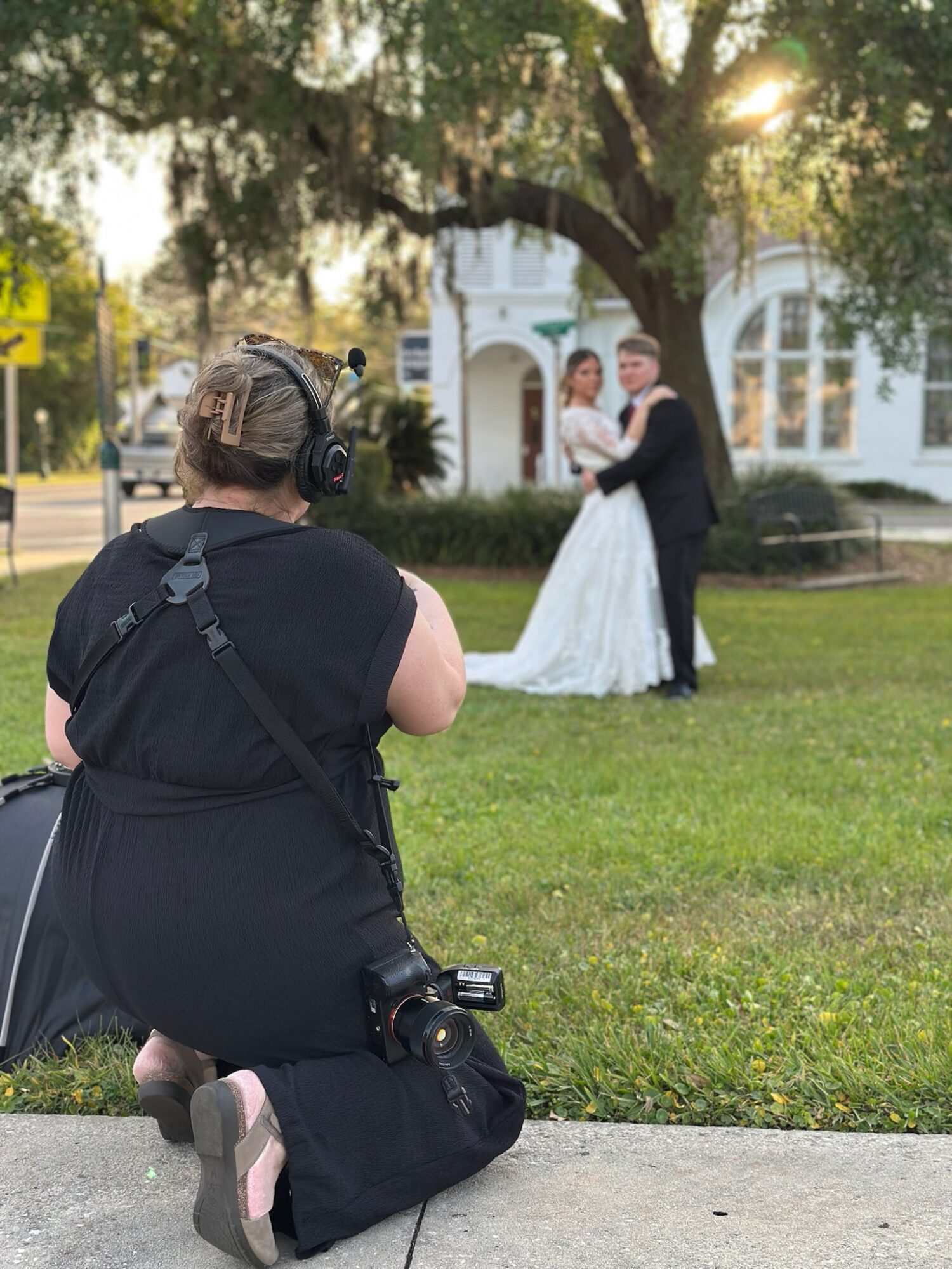 Photographer kneeling on grass taking picture of a bride and groom dancing outdoors, with trees and a building in background.