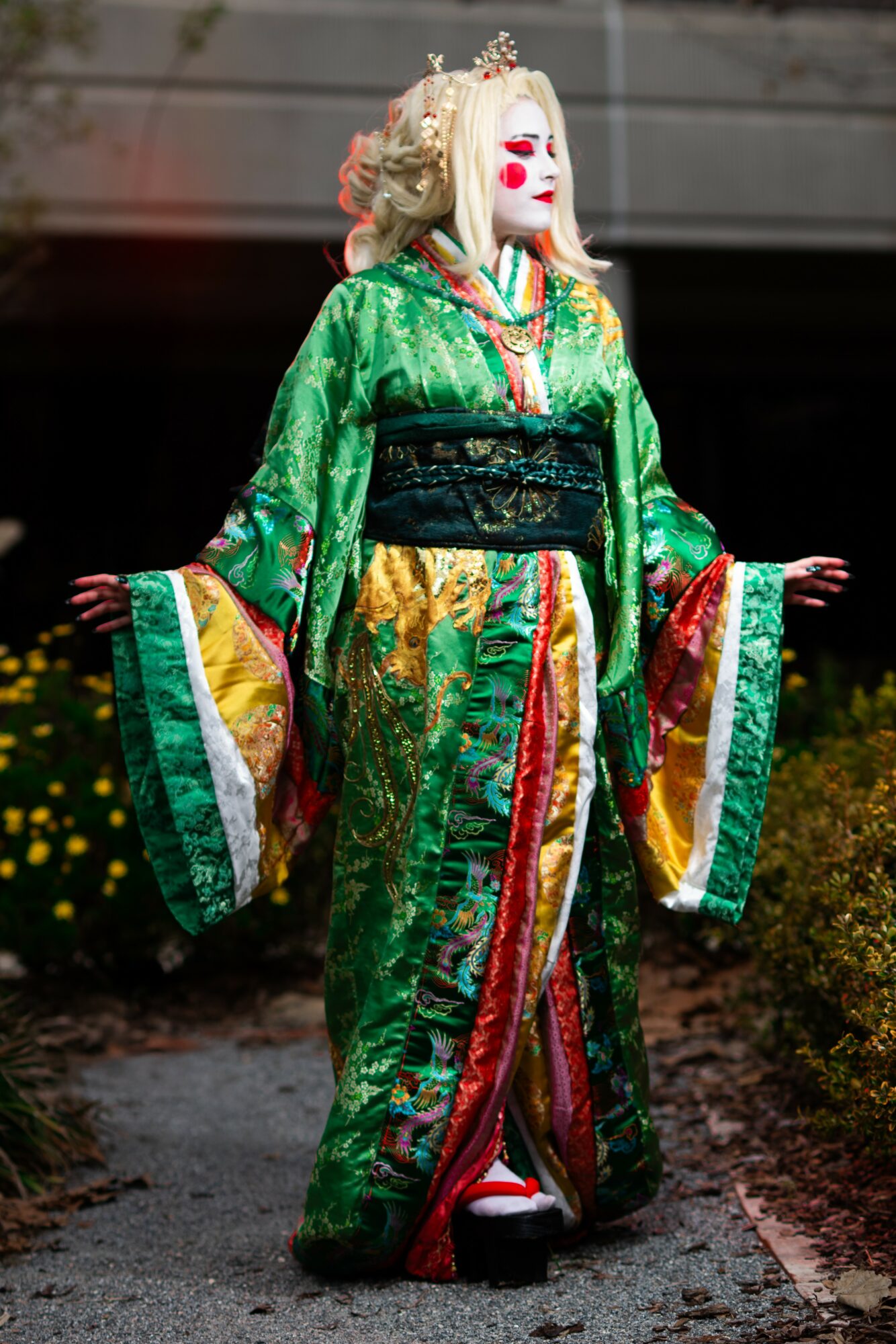 Person in traditional green kimono with colorful patterns, standing outdoors with arms extended, wearing a white mask and floral headpiece.