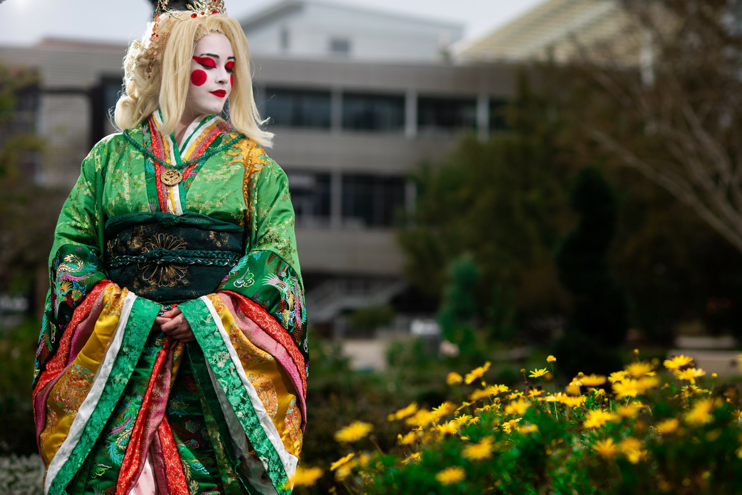 Person dressed as a clown with white face paint and red lips, wearing a colorful green, red, and gold costume, standing outdoors near yellow flowers.