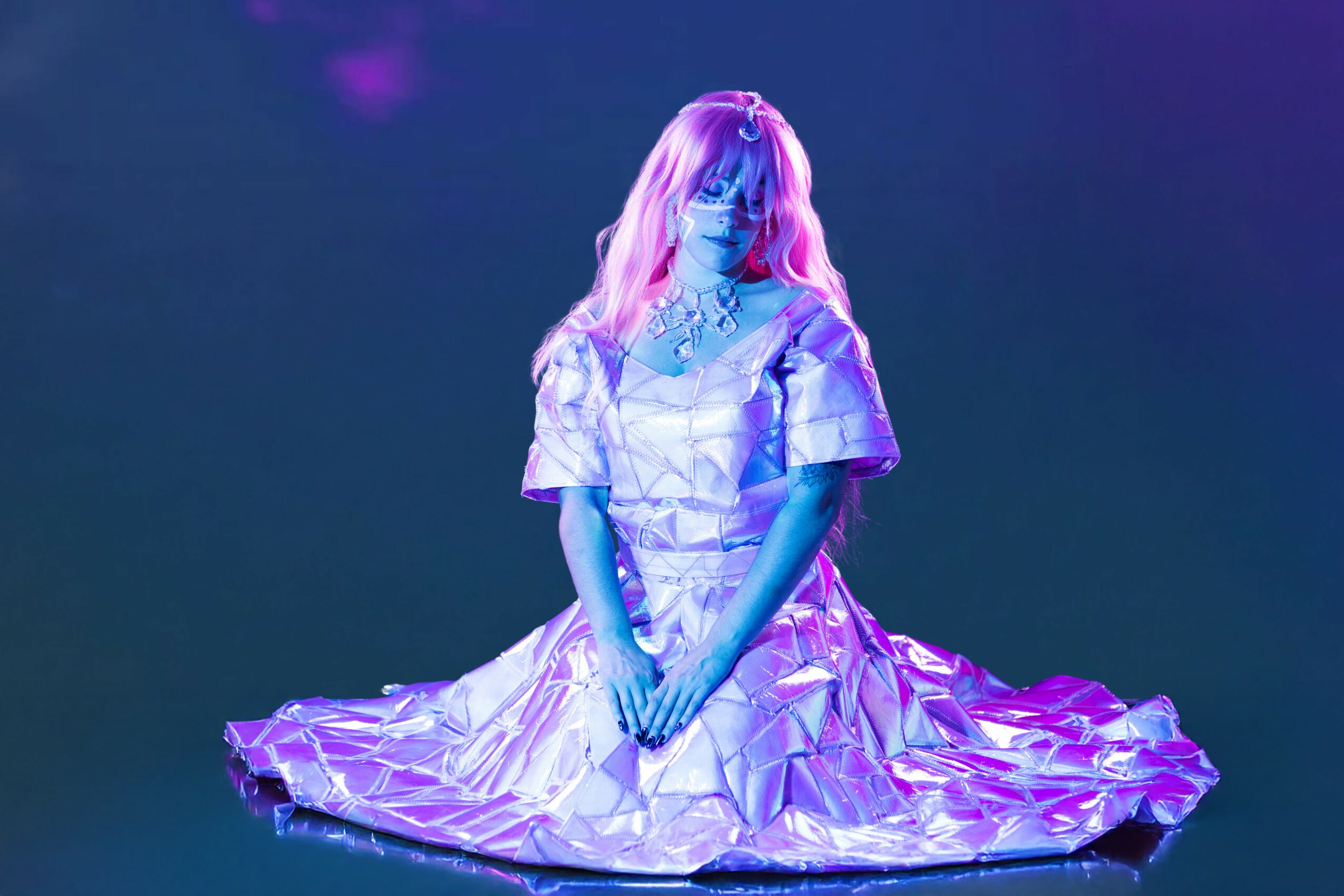 Person with long hair sitting on the floor, wearing a shiny, reflective dress, illuminated by purple and blue lighting.