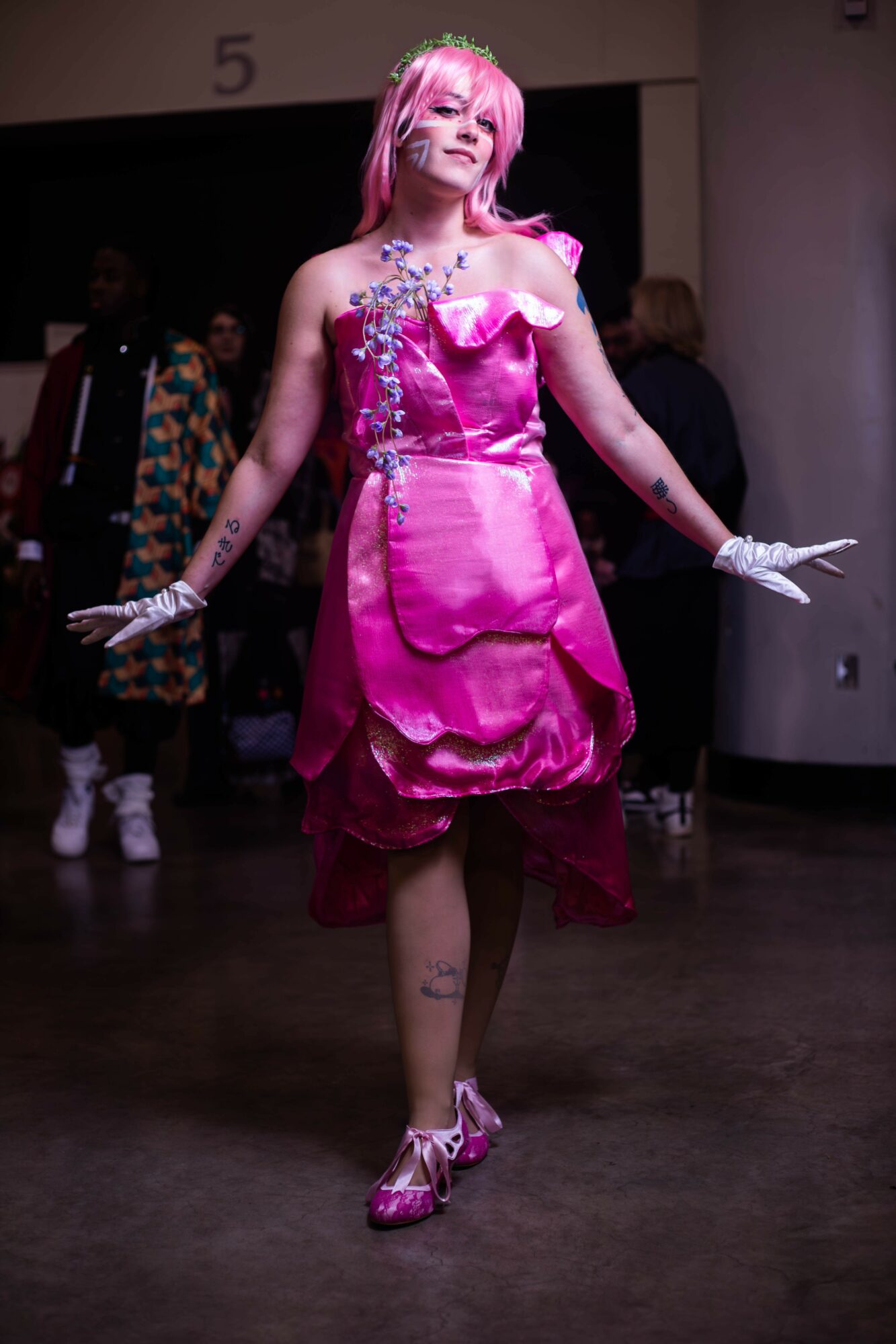 Person with pink hair in a shiny pink dress and gloves, walking indoors with people in the background.