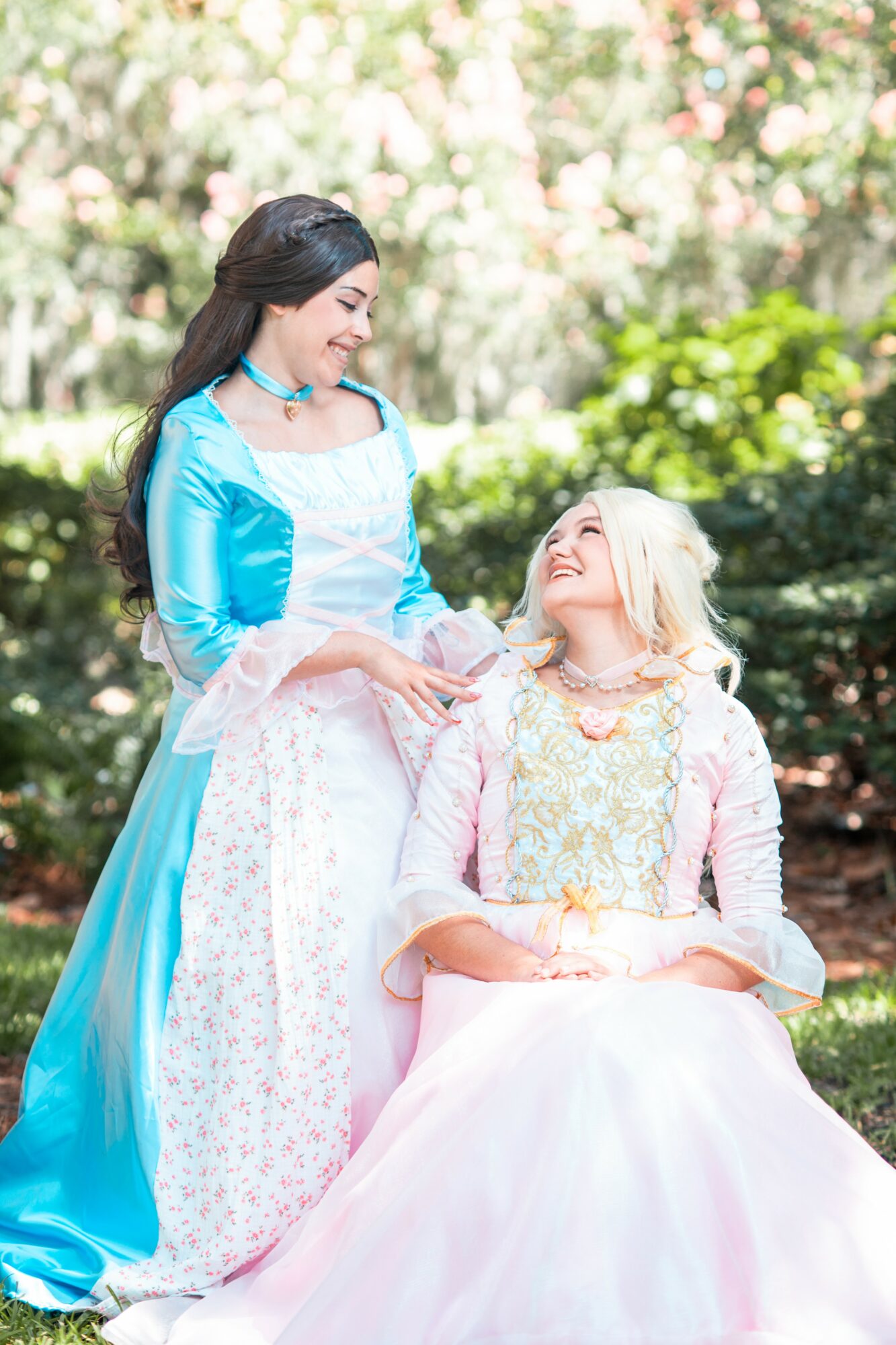 Two women in colorful dresses outdoors, one standing and smiling, the other sitting and looking up, surrounded by greenery.