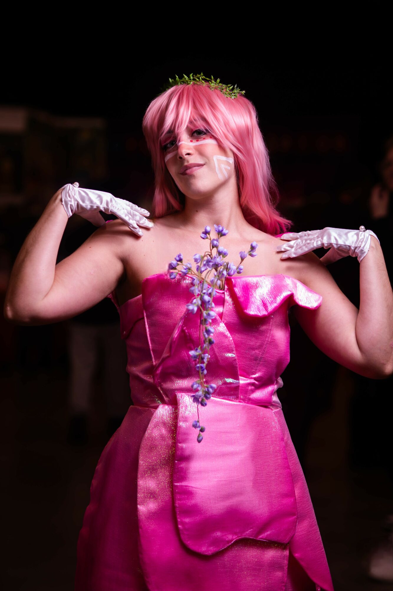 Person with pink hair in a pink dress holding a decorative chain, smiling, against a dark background.