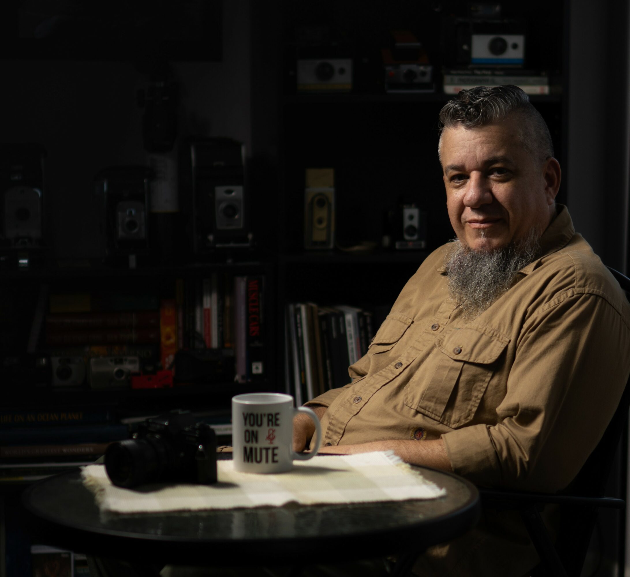 Man with a beard sitting at a table with a mug and a camera, bookshelf in background.