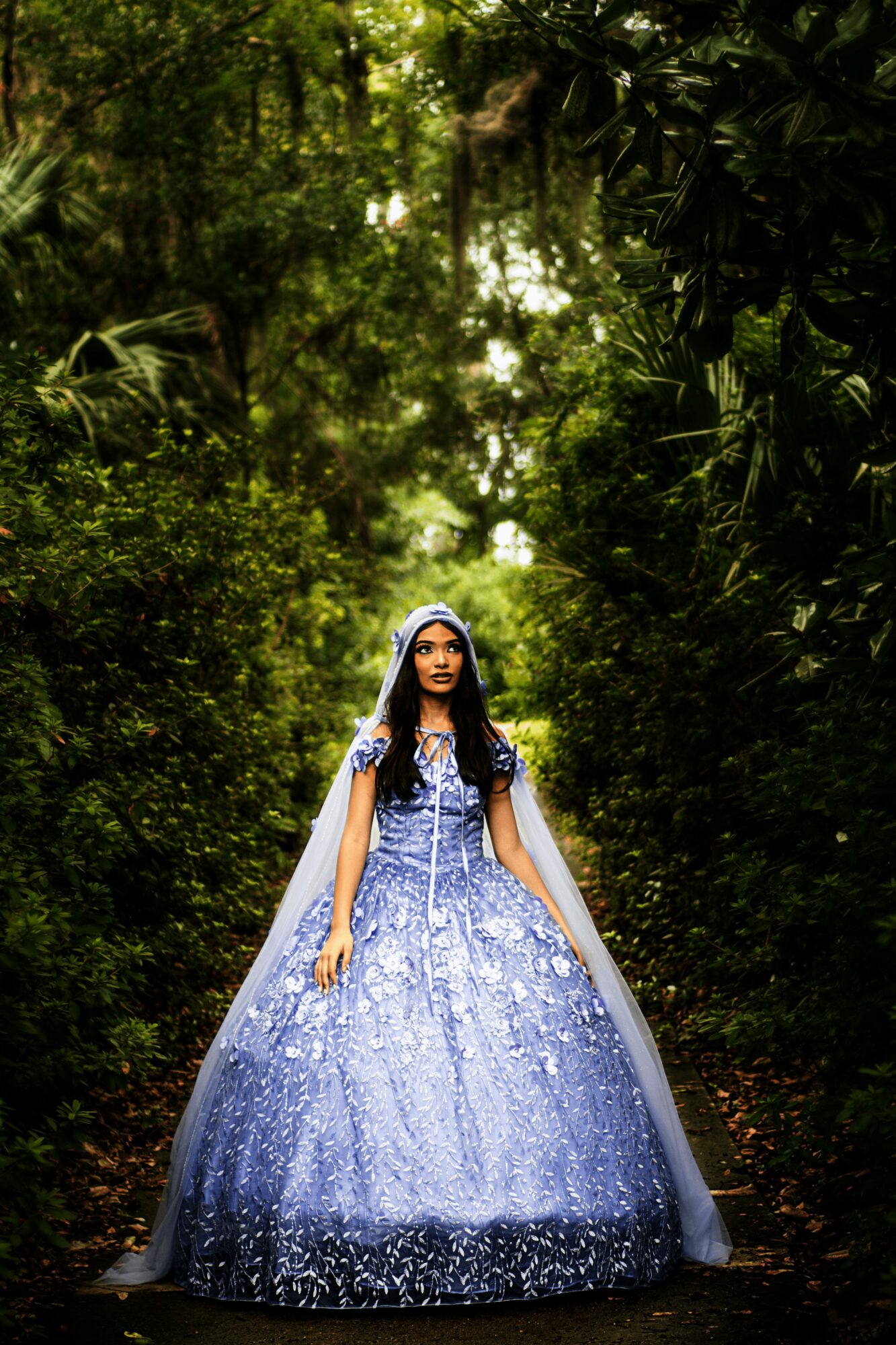 Woman in a blue and white gown with a cape standing in a lush green forest, sunlight filtering through trees.