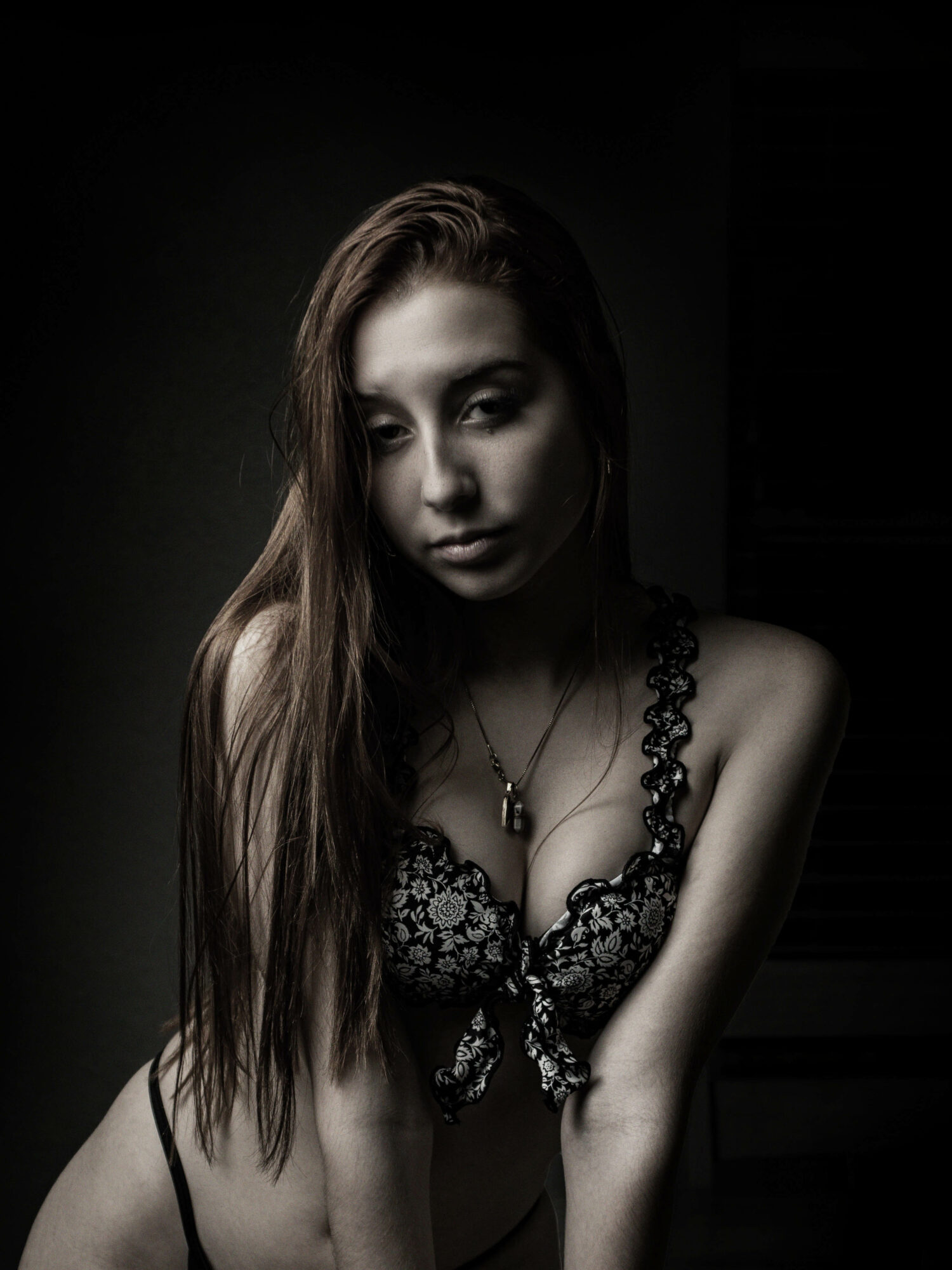 Young woman with long hair in a lace top, looking down, dim lighting, dark background.