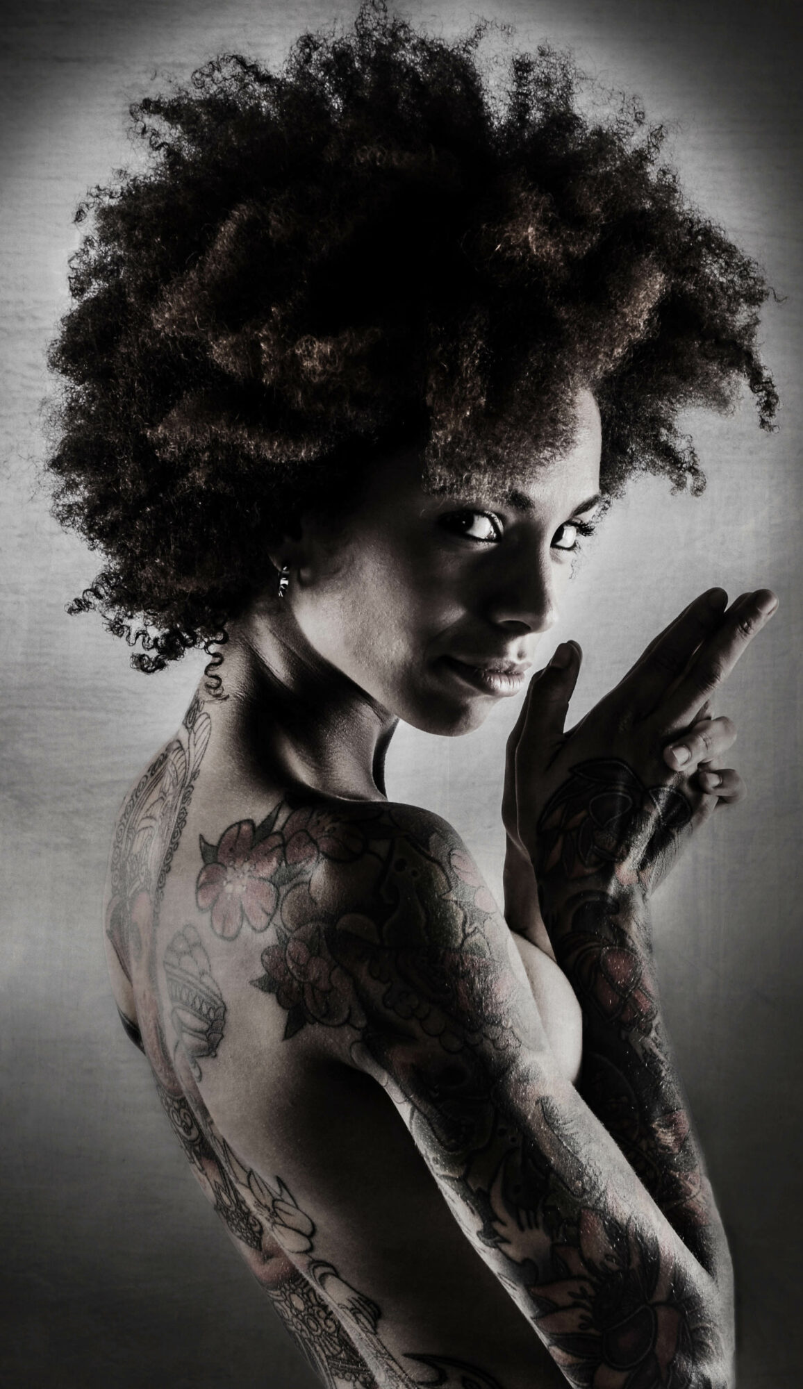 Black and white portrait of a woman with natural curly hair, tattoos on her arm, and a contemplative expression.