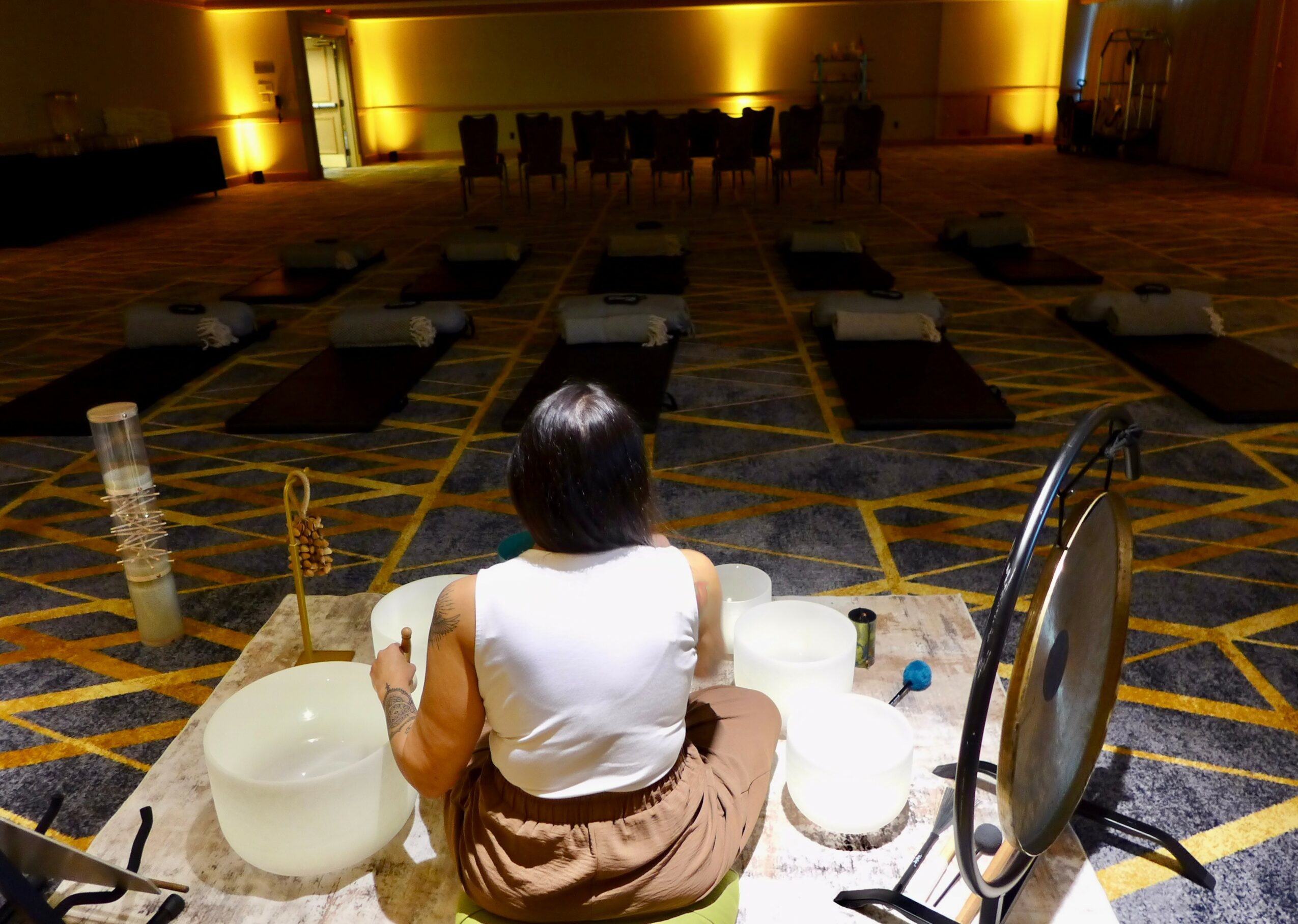 Person sitting on the floor with bowls and a mirror, facing a stage with chairs and yellow lighting.