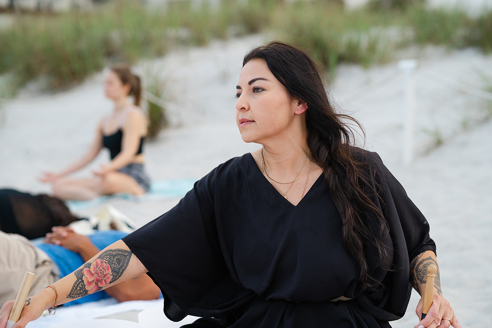 Woman with long dark hair and tattoos on arms sitting outdoors, with a blurred person in the background, on a sandy surface.