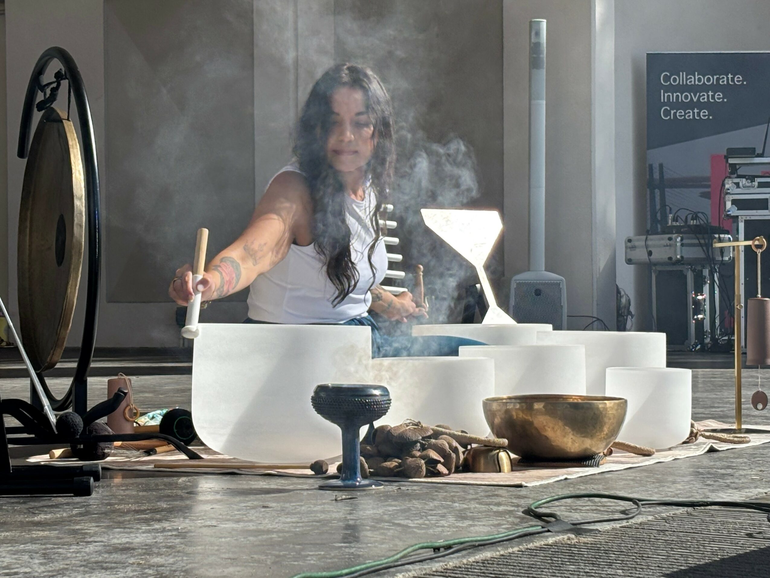Woman playing singing bowls with steam rising, surrounded by bowls and objects, in a modern room.