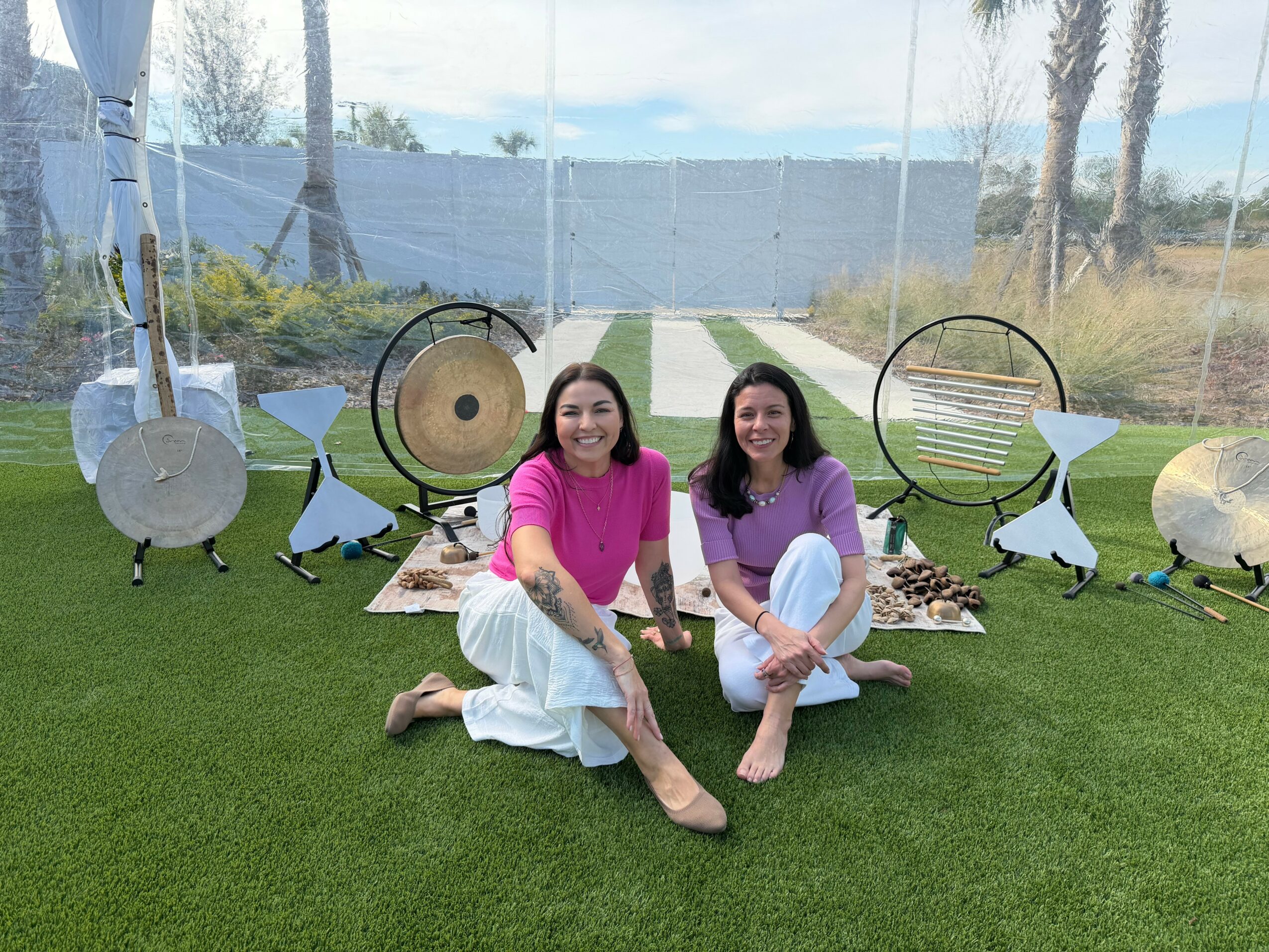 Two women sit on grass in front of musical instruments, smiling, with a glass wall and outdoor scenery behind them.
