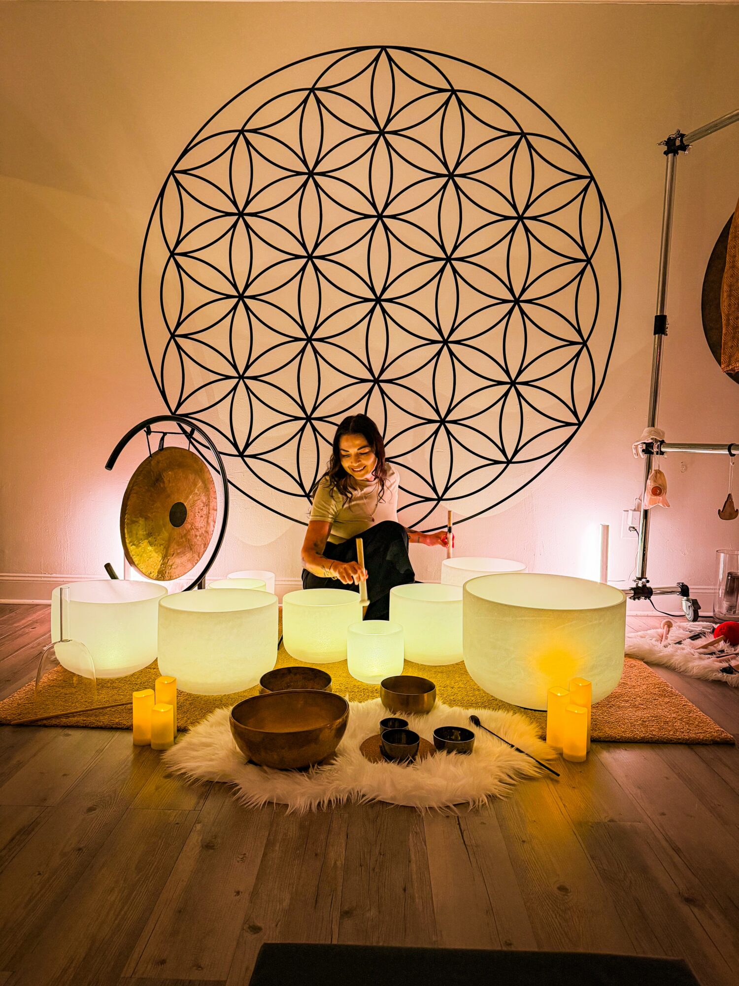 Person sitting on illuminated crystal bowls with candles, in a room with a large geometric wall art, musical instruments, and candles.