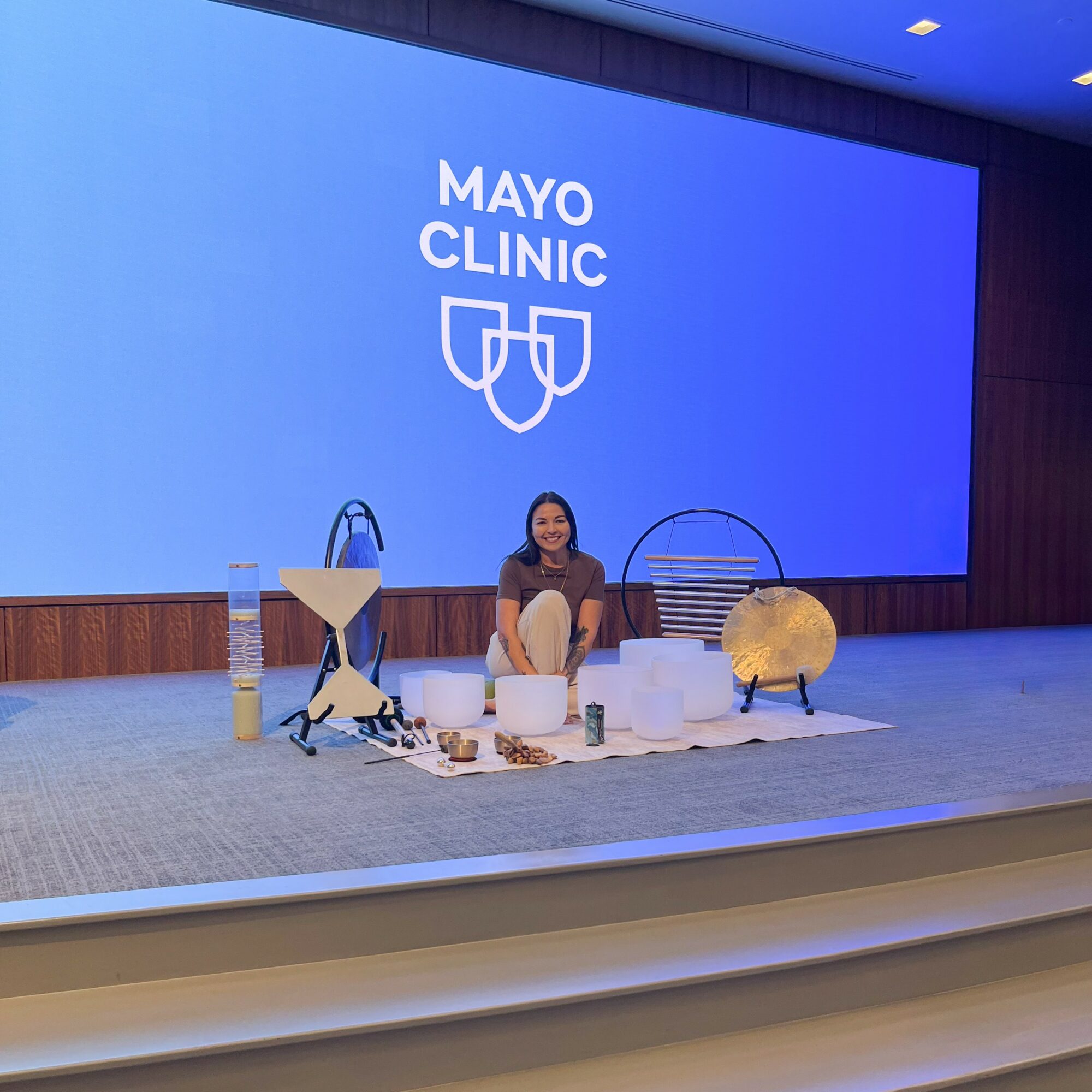 Person sitting on stage with medical equipment and supplies, large blue screen behind displaying Mayo Clinic logo.