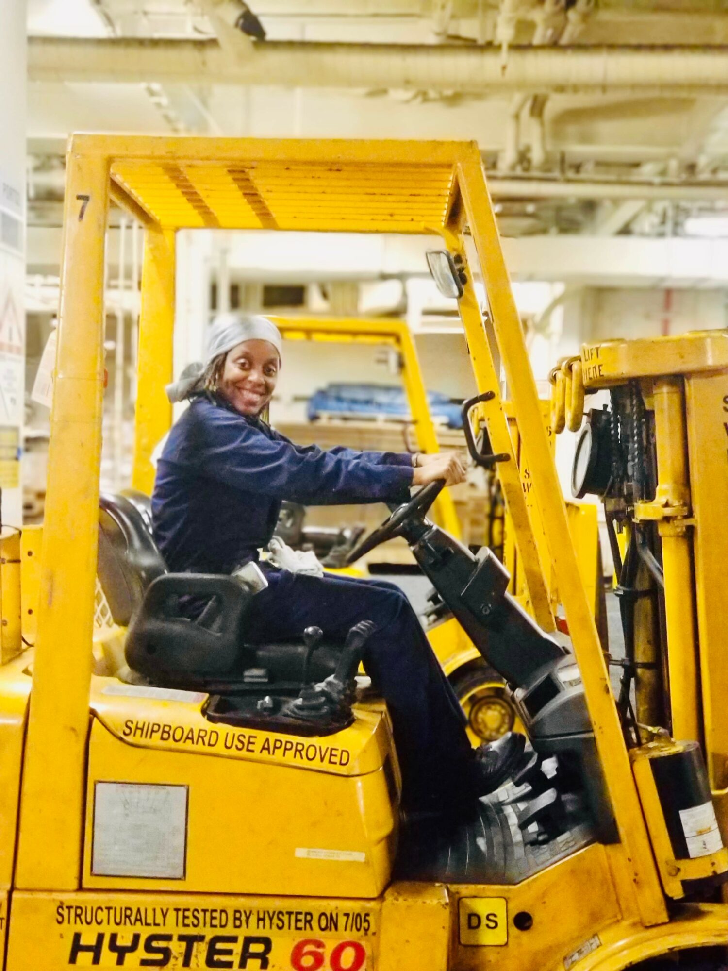 Woman operating a yellow forklift inside a warehouse, smiling, with industrial equipment in background.