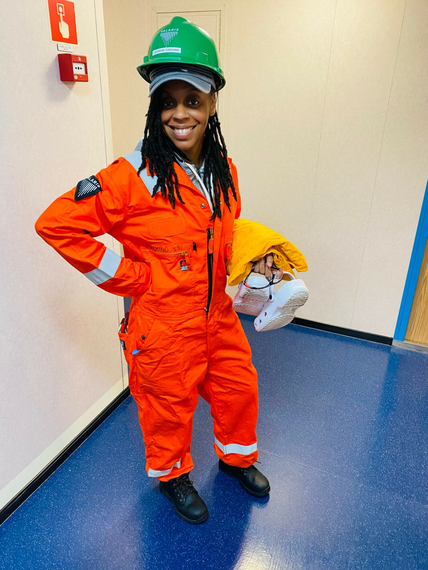 Person in orange coveralls and green helmet holding yellow jacket, standing indoors with blue floor and beige wall.