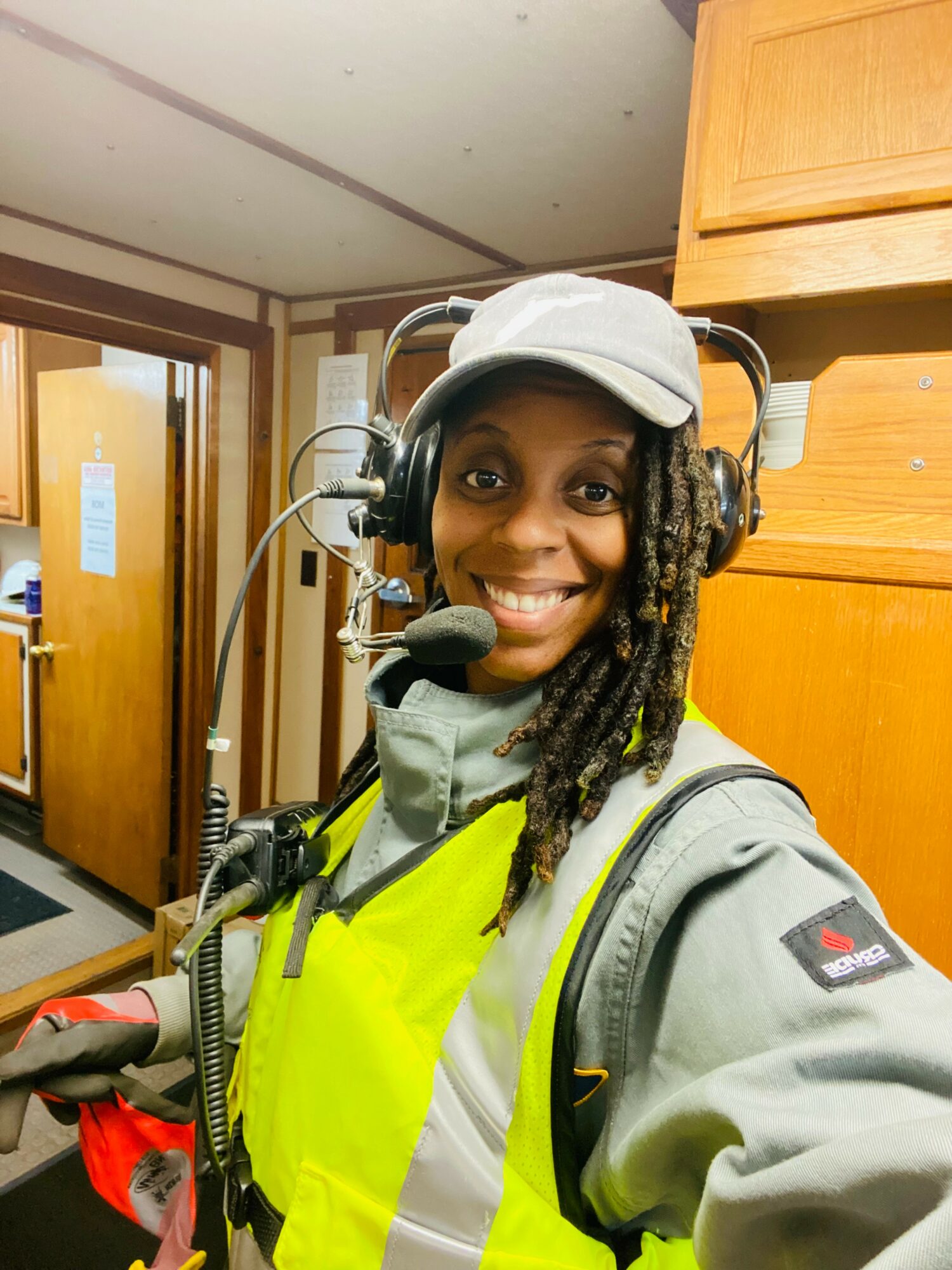 Smiling person wearing a headset, gray cap, and high-visibility vest indoors, with wooden cabinets in the background.