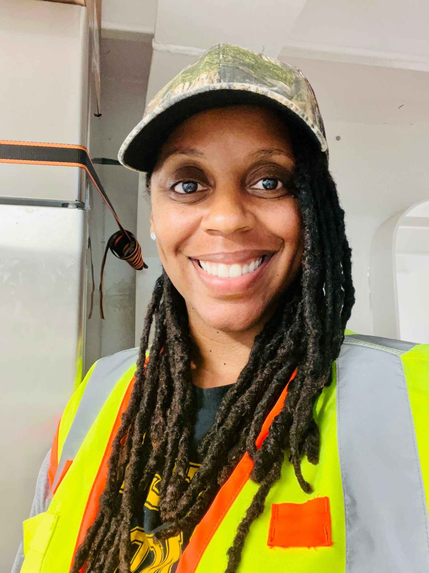 Smiling woman wearing a camouflage cap and high-visibility vest indoors.