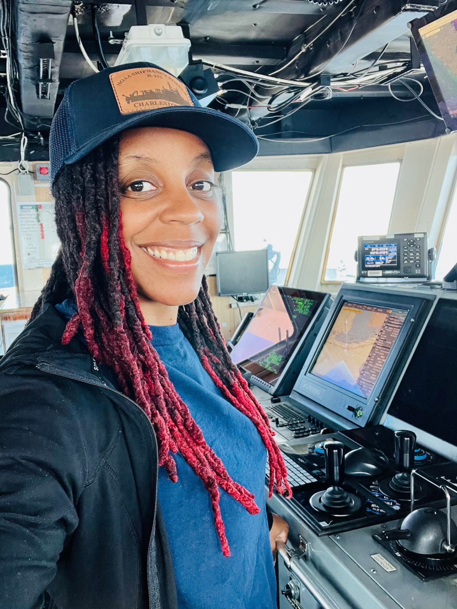 Smiling woman with red dreadlocks wearing a cap, inside a control room with multiple screens and equipment.