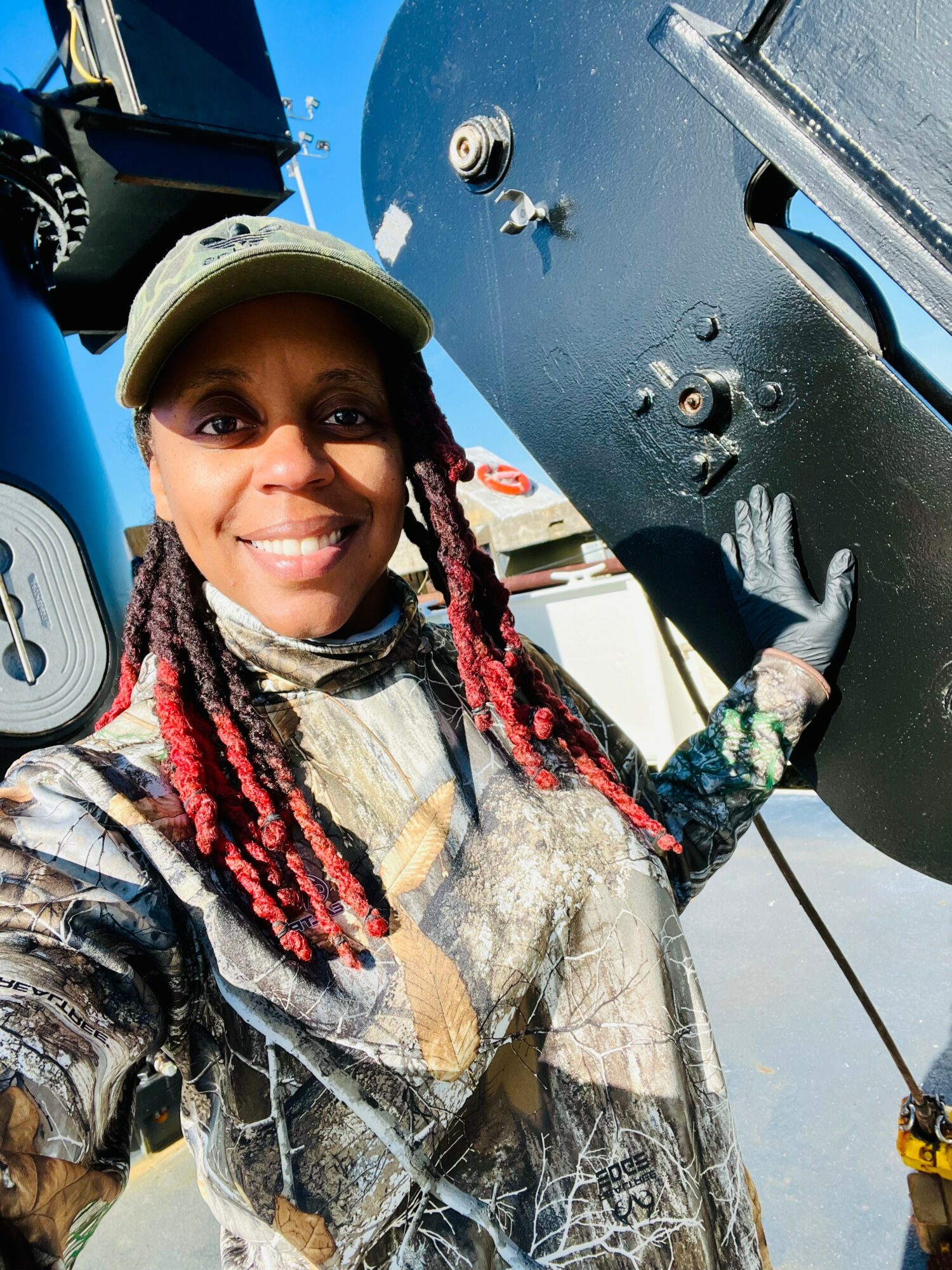 Smiling woman in camouflage clothing and cap outdoors, holding a large black object, with blue sky in background.