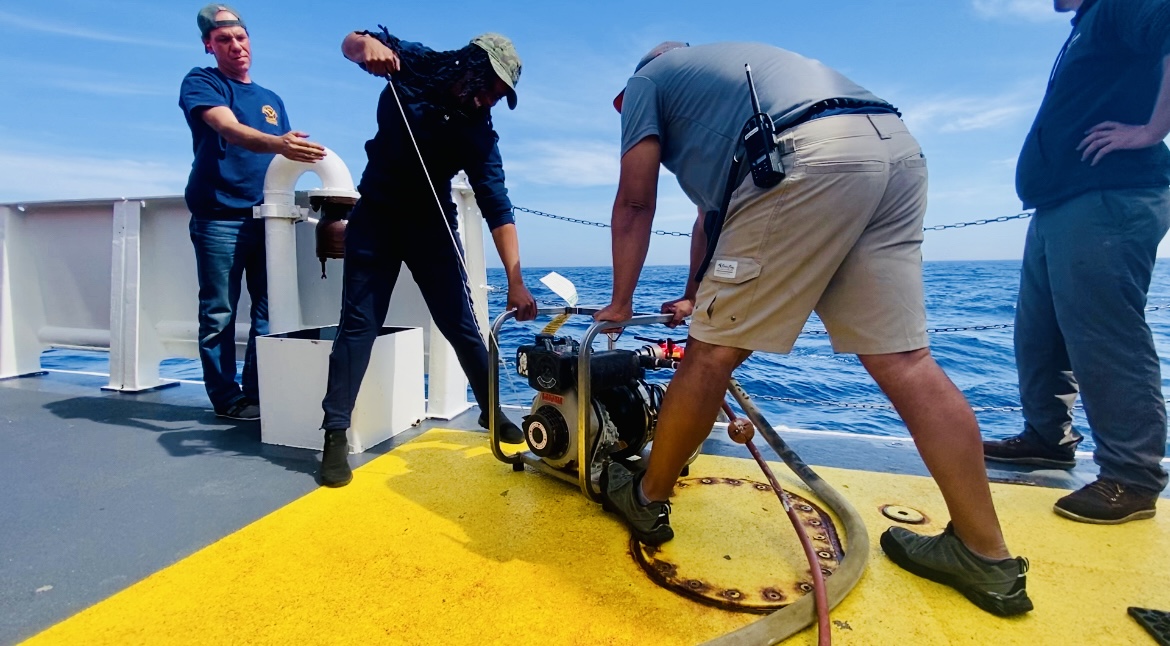 Four people on a boat deck with equipment, one person operating a device, ocean in background, clear sky.