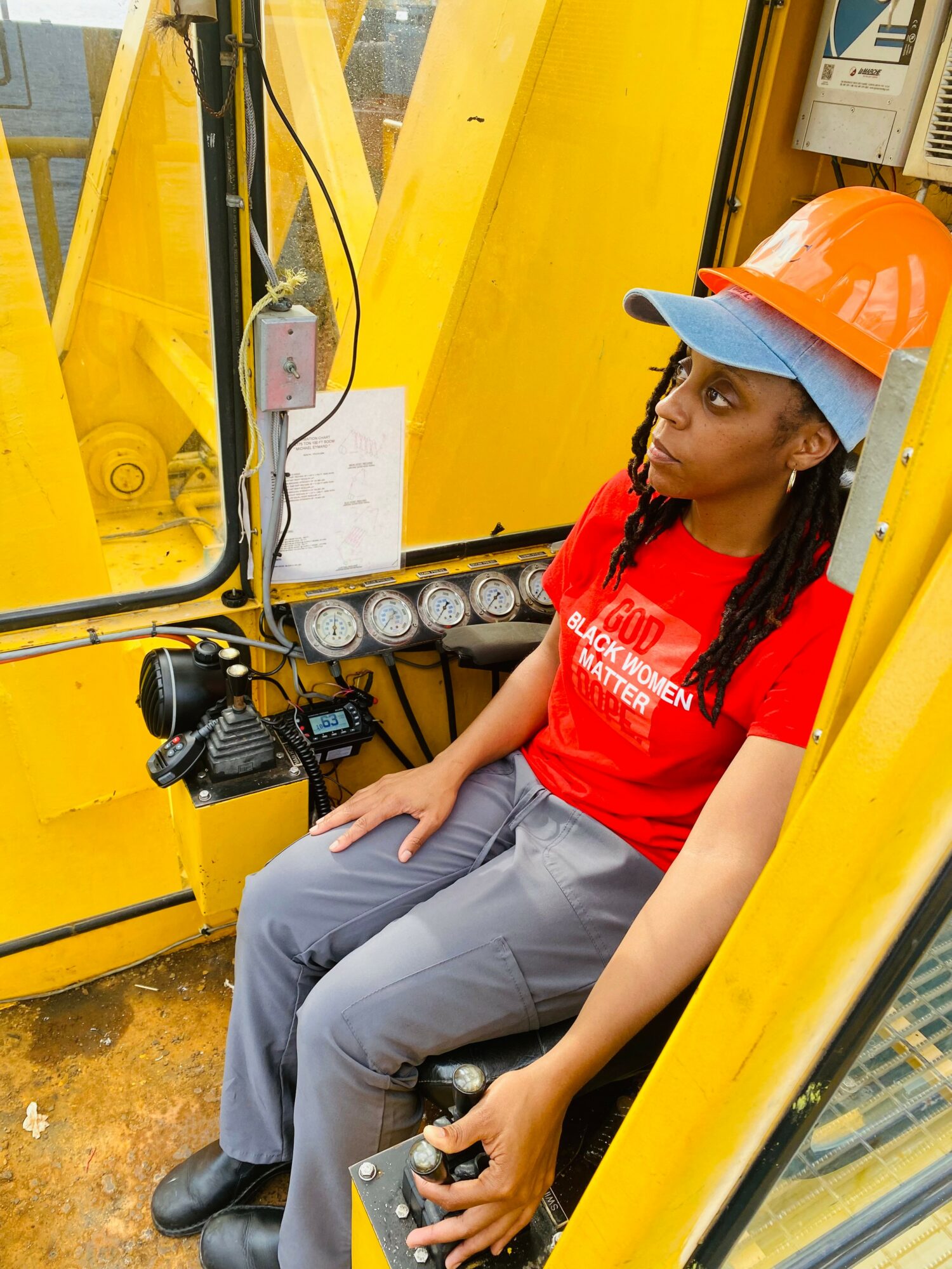 Woman wearing an orange safety helmet and red shirt sitting inside industrial equipment, holding a control device.