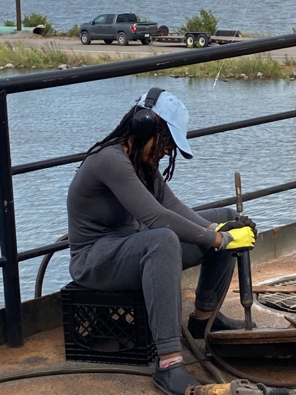 Person sitting on a boat, wearing a gray hoodie, yellow gloves, and a white cap, holding a fishing rod near water.