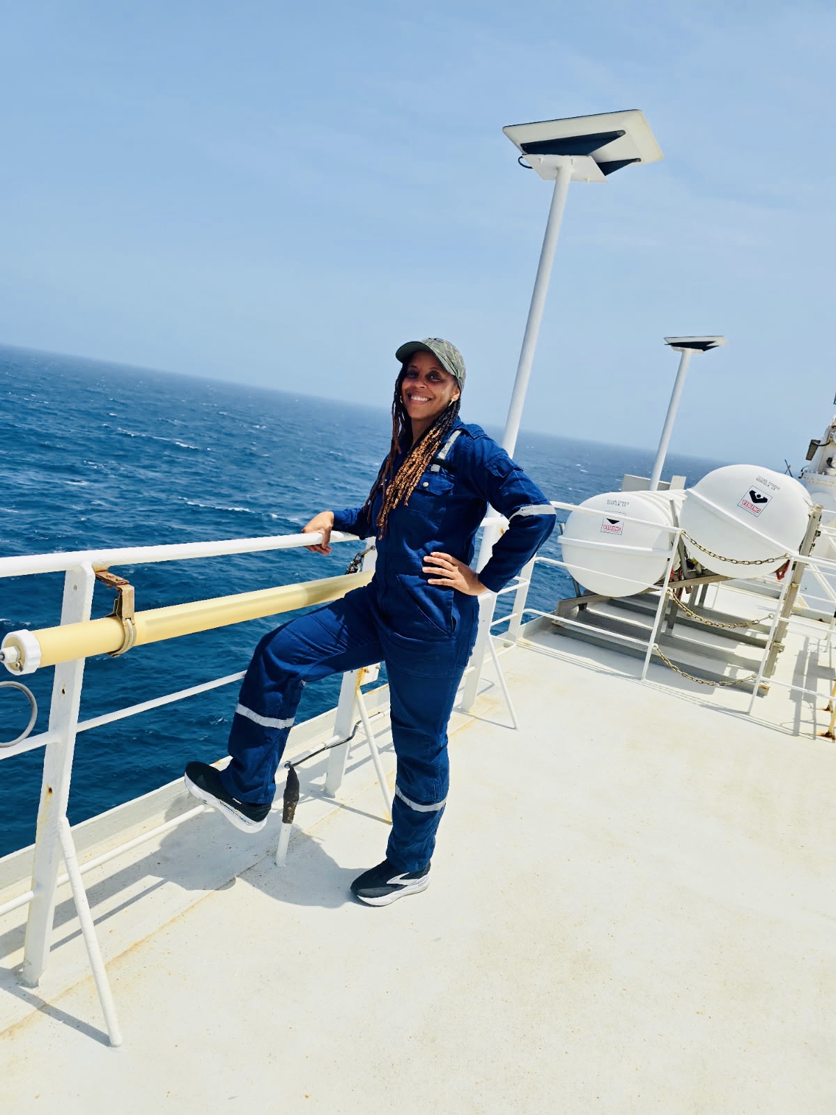 Woman in blue jumpsuit and hat standing on ship deck with ocean background, smiling, with equipment behind her.