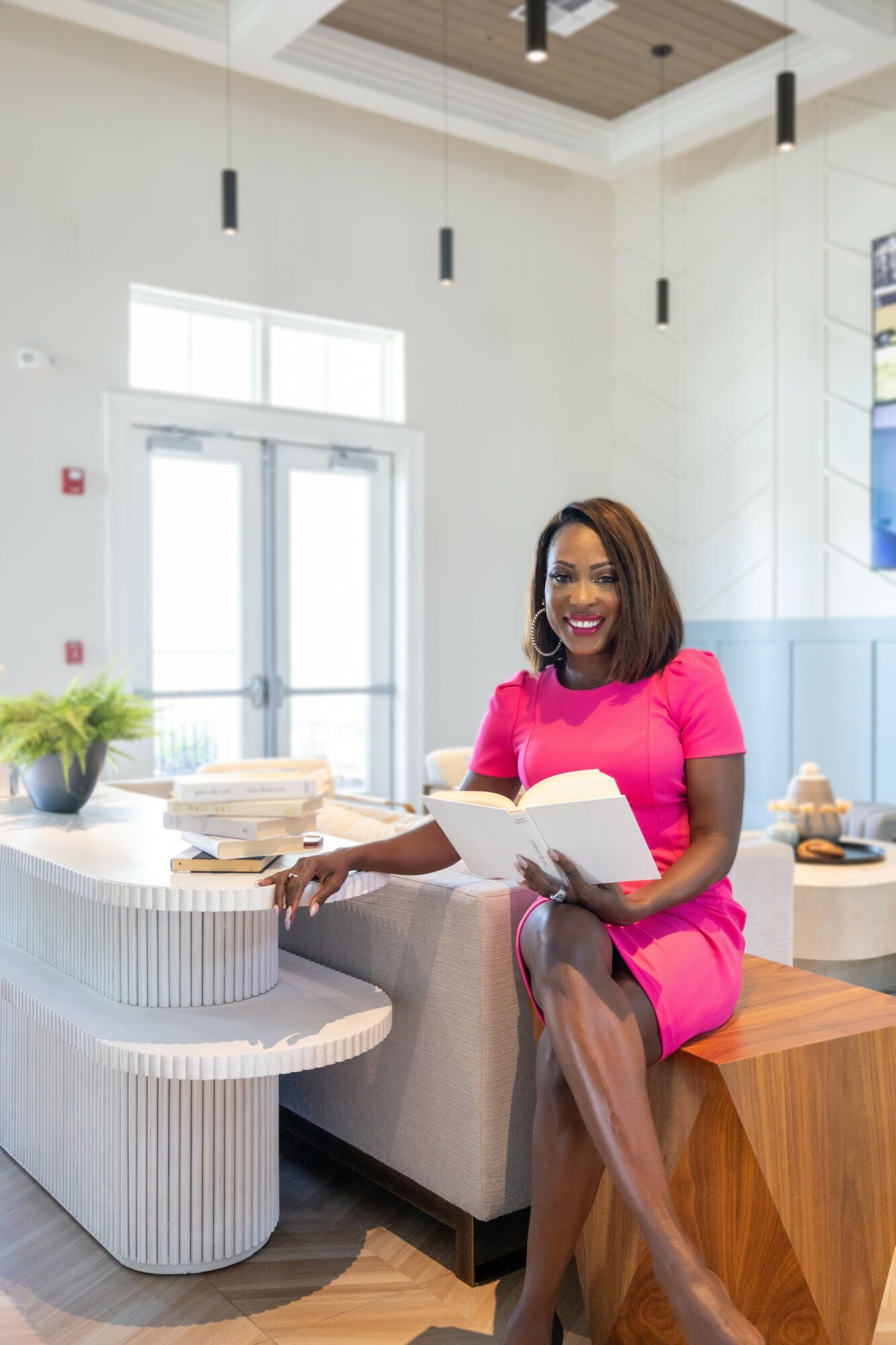 Smiling woman in pink dress sitting on a wooden bench, reading a book in a bright, modern room with large windows.