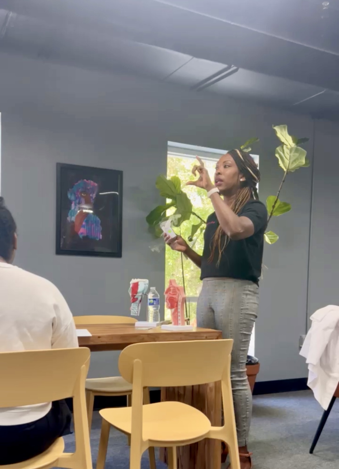 Woman standing and speaking in a room with people seated, plants, and a framed picture on the wall.