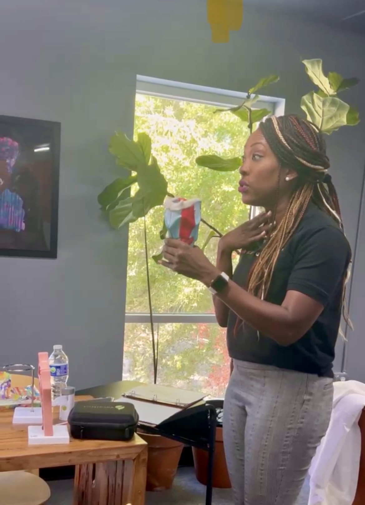 Woman with braided hair holding a colorful cup in a room with a window and green plants, sitting near a table.