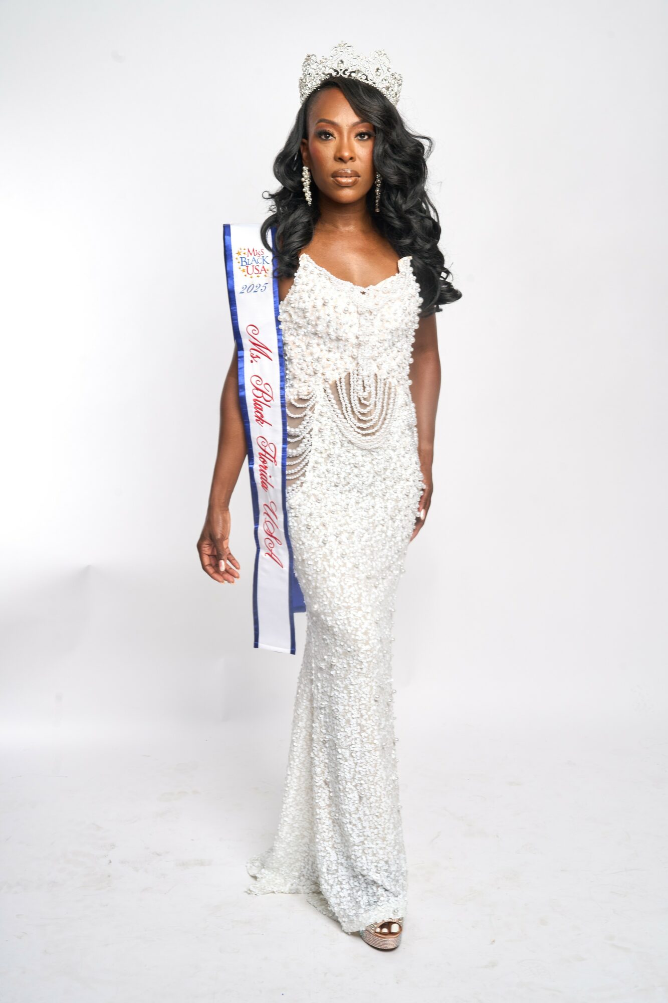 Woman in white gown with crown and sash, standing against a white background.