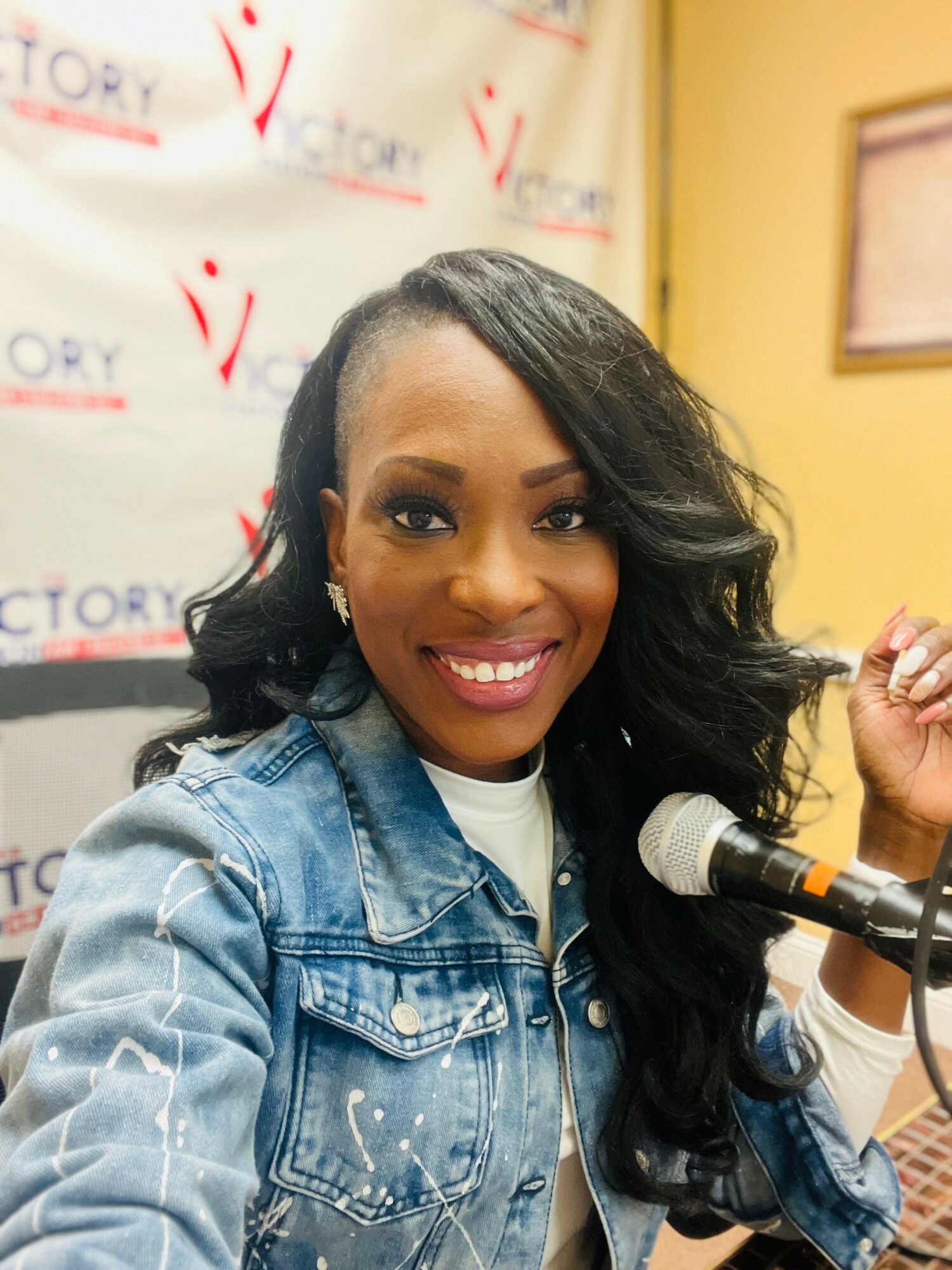 Smiling woman with long, wavy black hair wearing a denim jacket, sitting at a microphone in a room with a yellow wall and a banner in the background.
