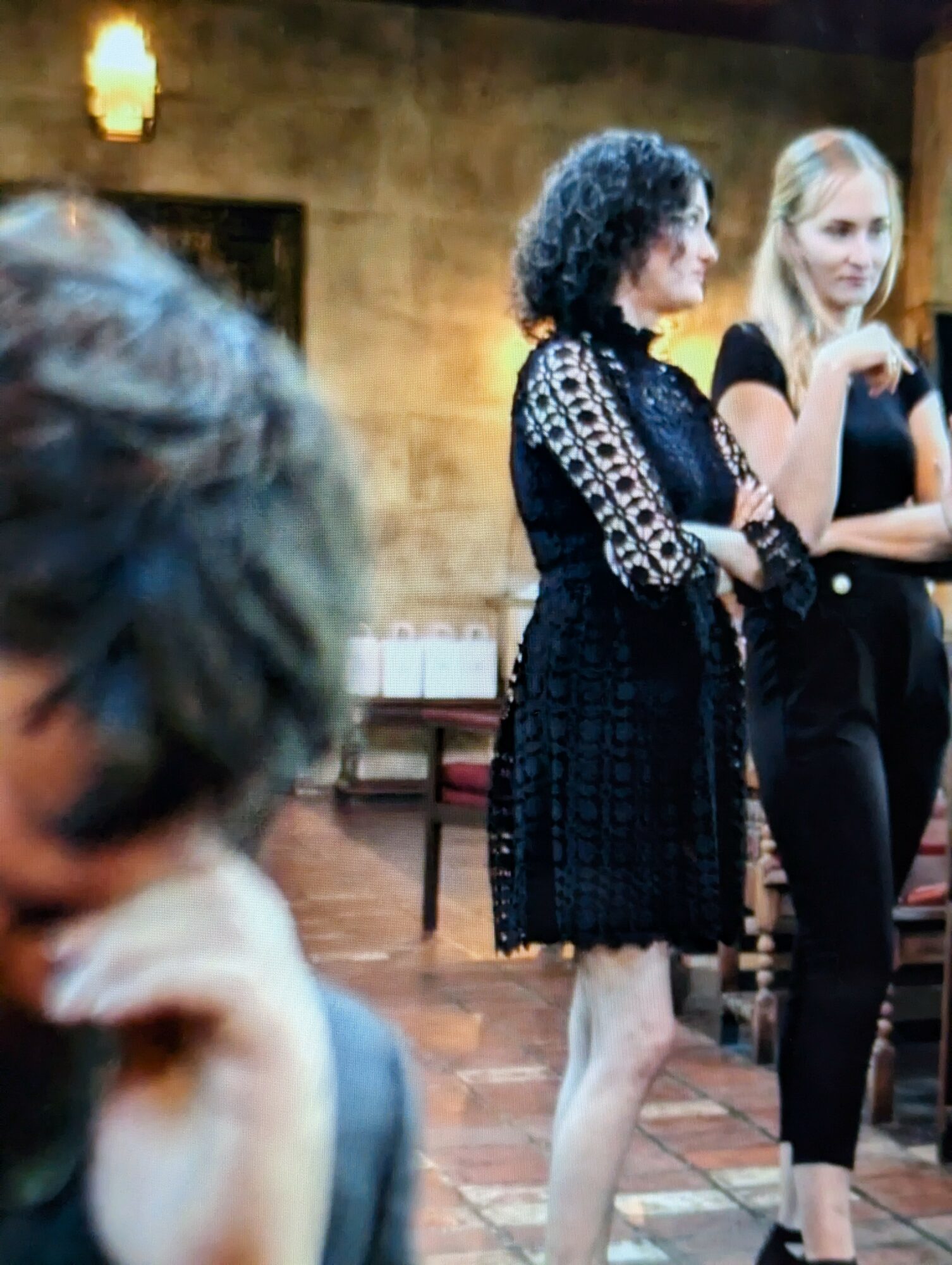 Two women standing and talking in a room with warm lighting and a textured wall, one with curly hair and the other with straight blonde hair.