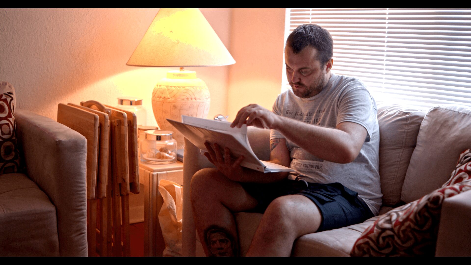 Man sitting on a sofa reading a magazine in a cozy living room with a lamp and window blinds