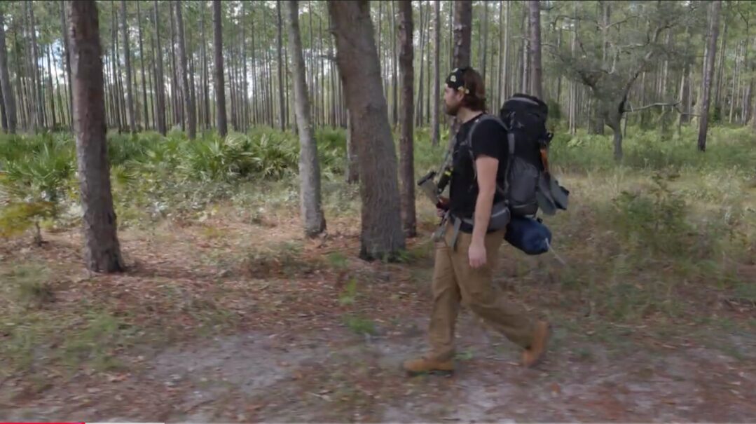 Person walking in a forest with a large backpack, trees and greenery around, walking on a dirt path.