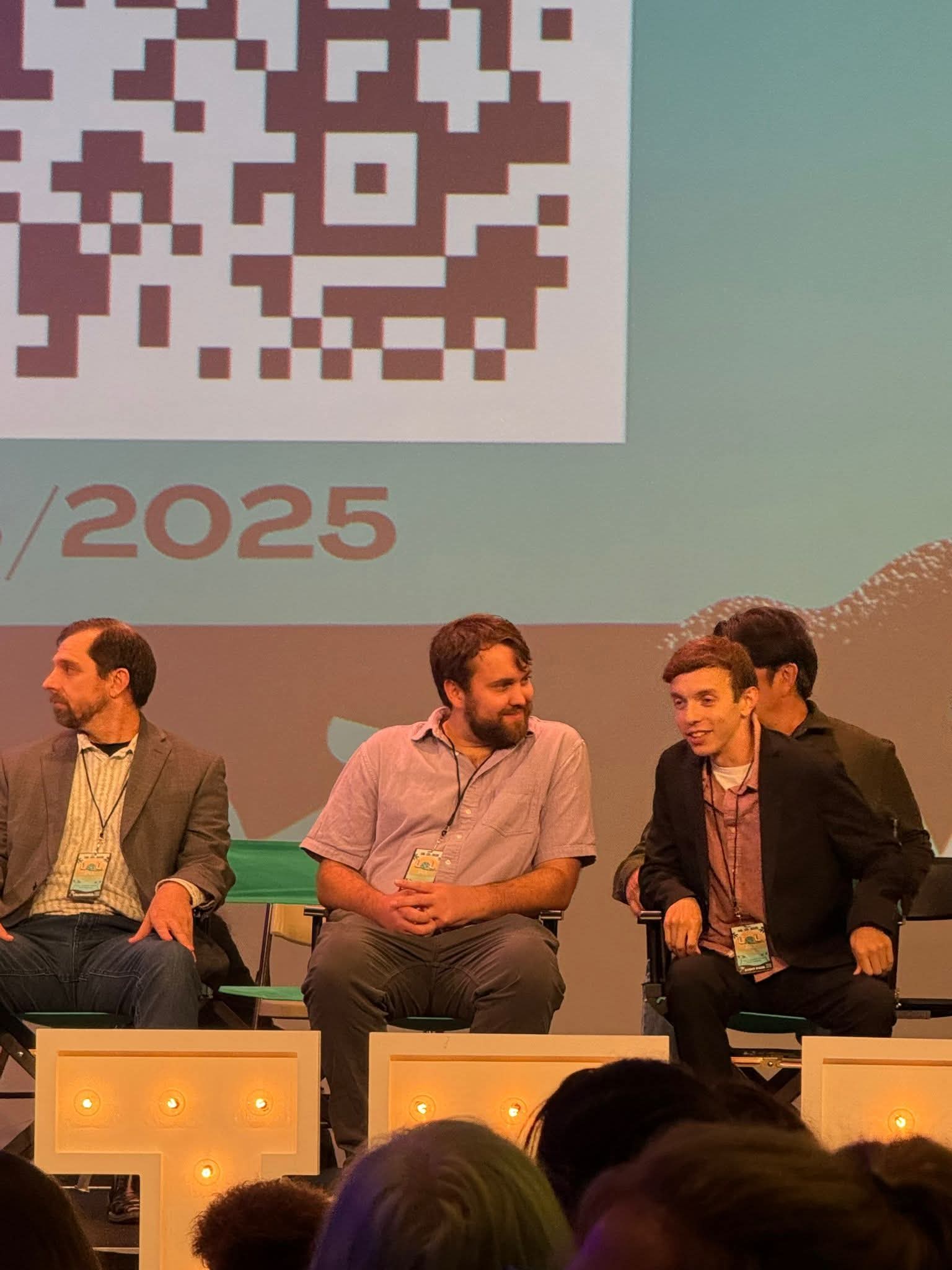 Three men sitting on a stage, engaged in conversation, with a large screen behind them displaying a QR code and the year 2025.