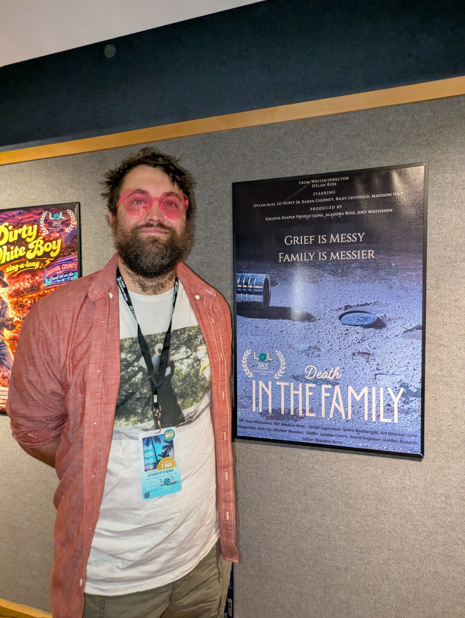 Man with glasses and beard wearing a lanyard, standing next to a movie poster on a wall.