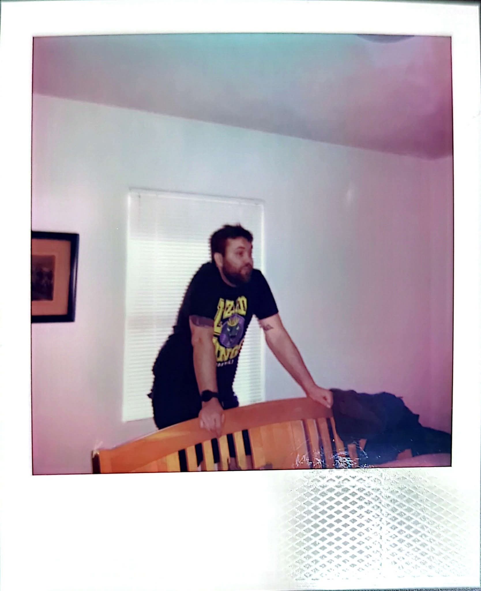 Man leaning on a wooden chair in a room with a window and framed picture on the wall.