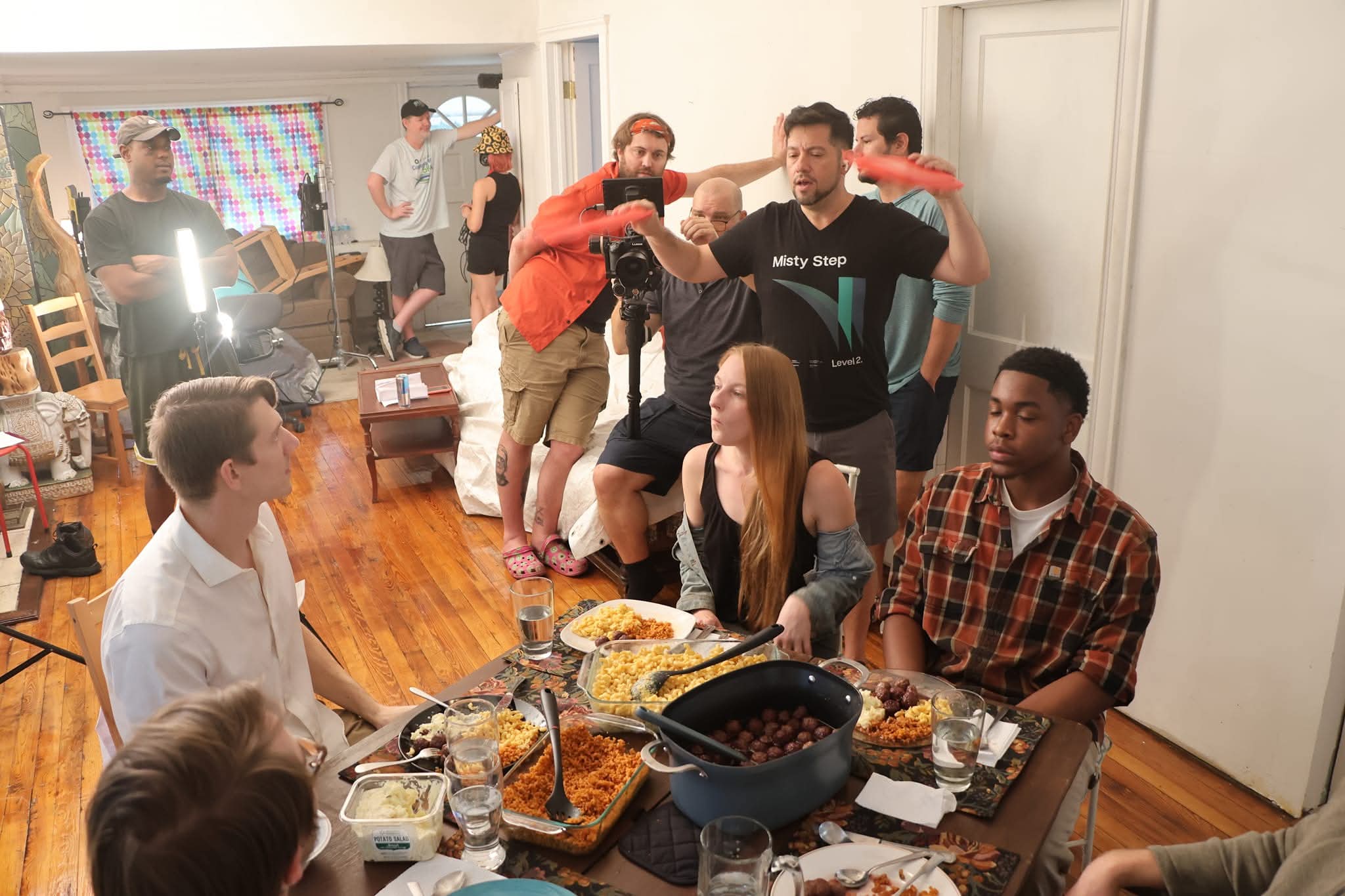 Group of people gathered around a table with food, some standing and some sitting, in a room with wooden floors.