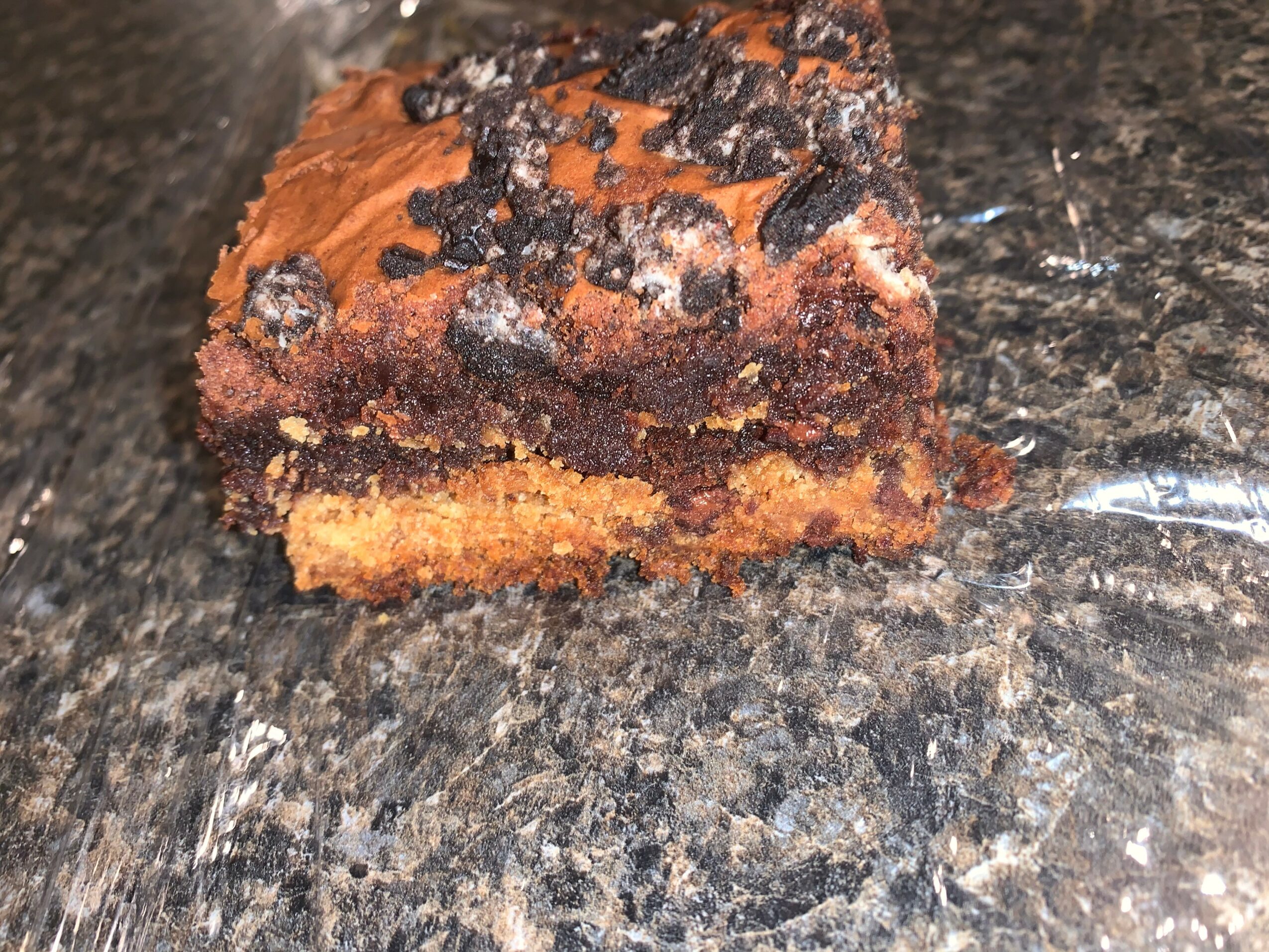 A piece of chocolate chip cookie cake on a dark granite countertop, showing a layered texture with chocolate chips on top.