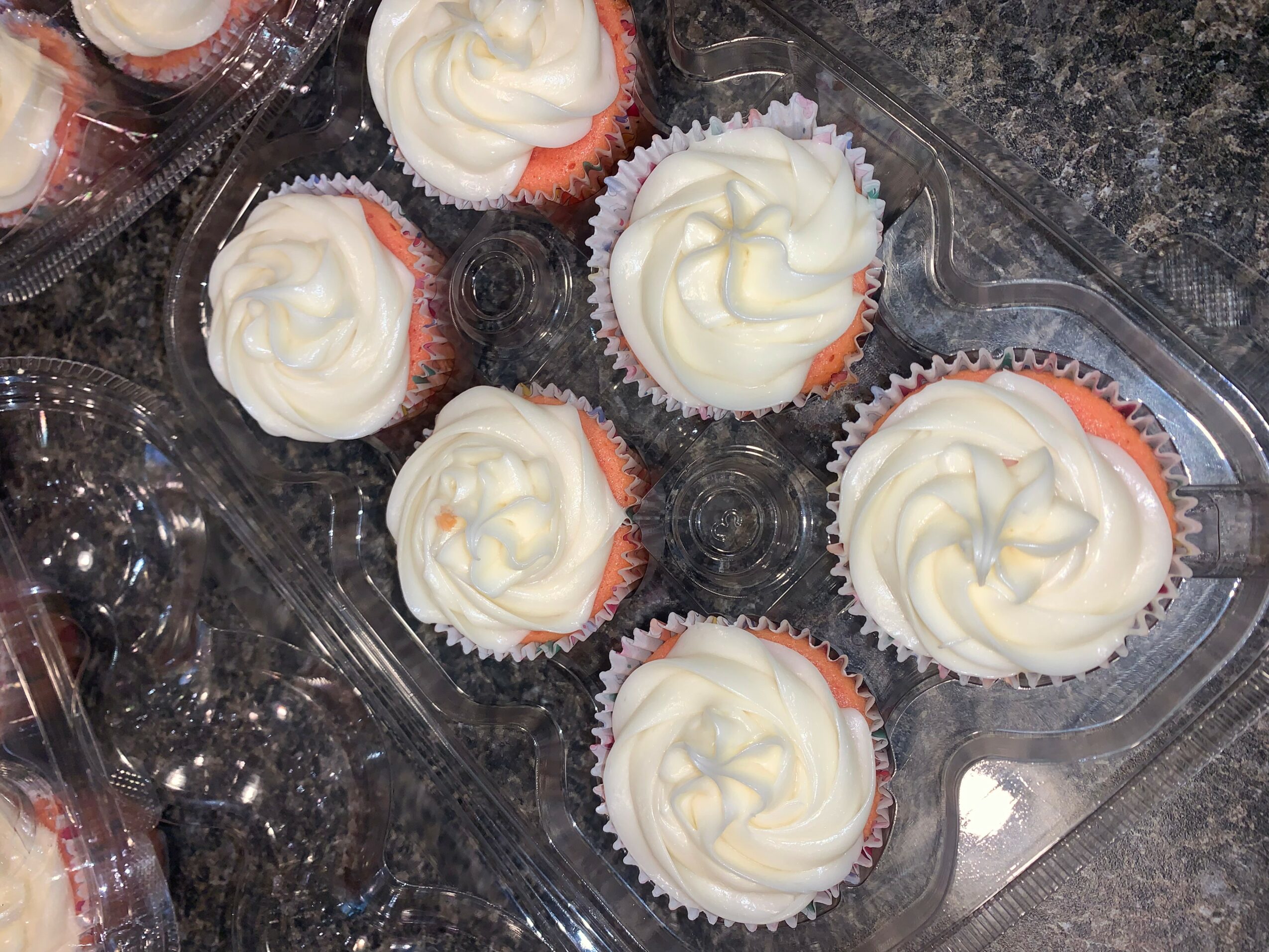 Cupcakes with white frosting in a plastic container on a dark surface.