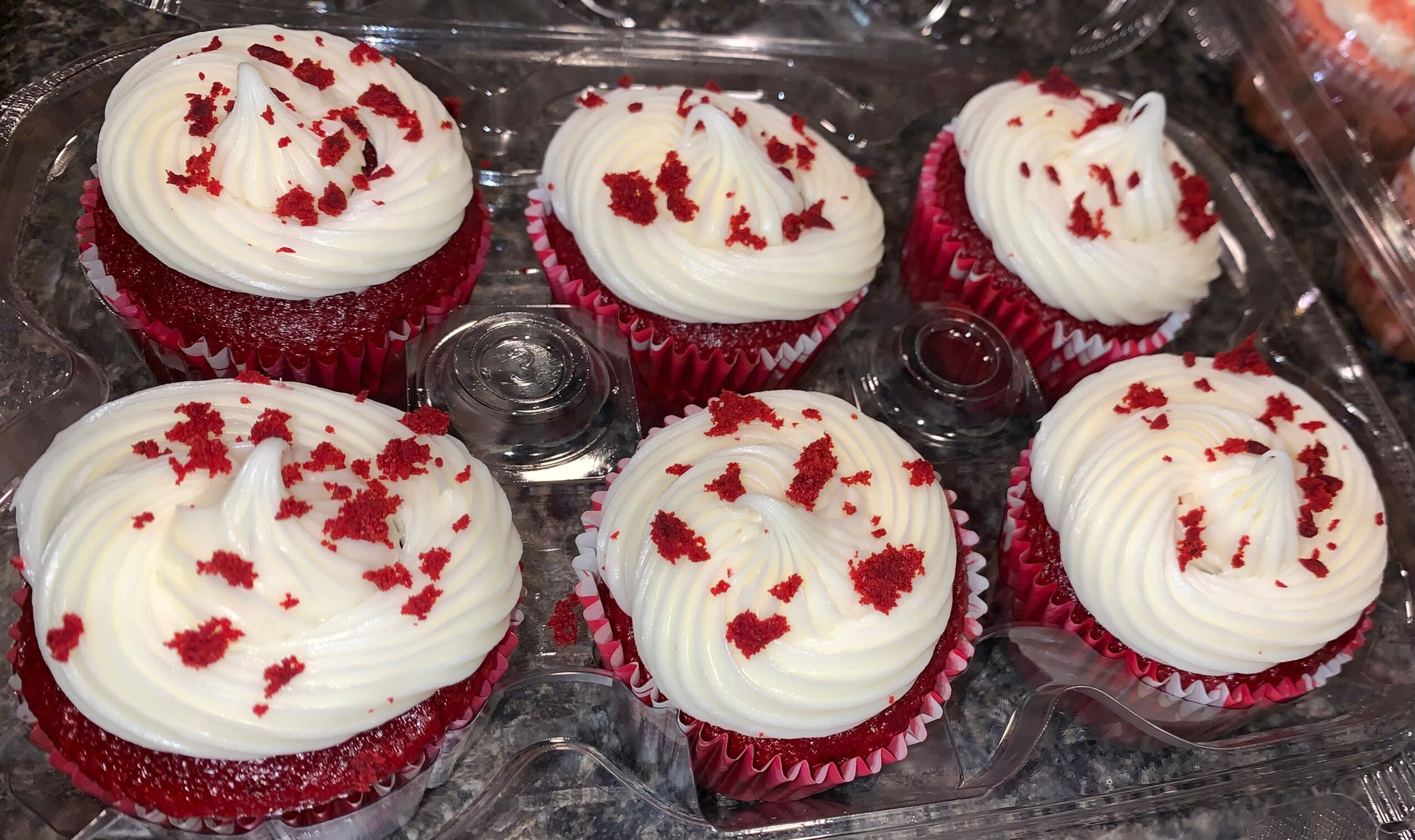 Six red velvet cupcakes with white frosting and red sprinkles in a plastic container.