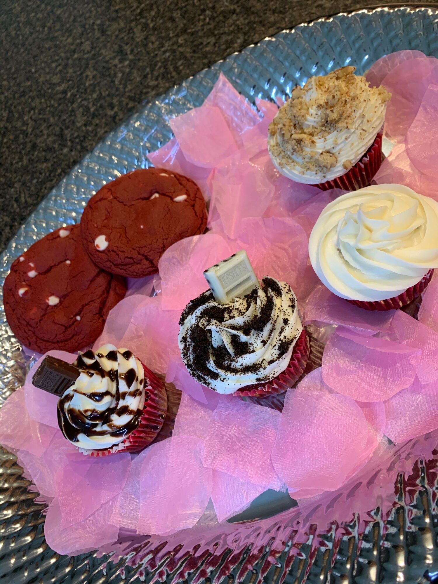 Six cupcakes on pink tissue paper in a silver tray, with various toppings and decorations.