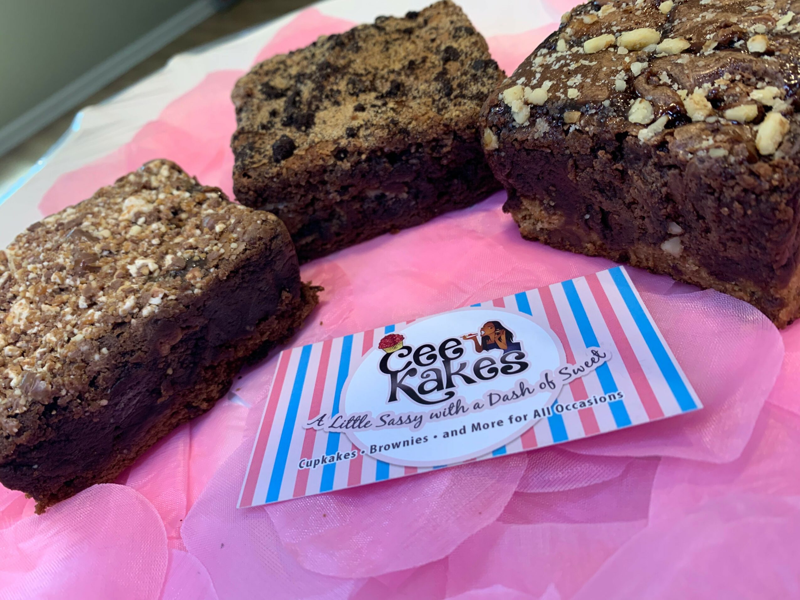 Three chocolate brownies on pink tissue paper with a striped bakery business card, close-up view.