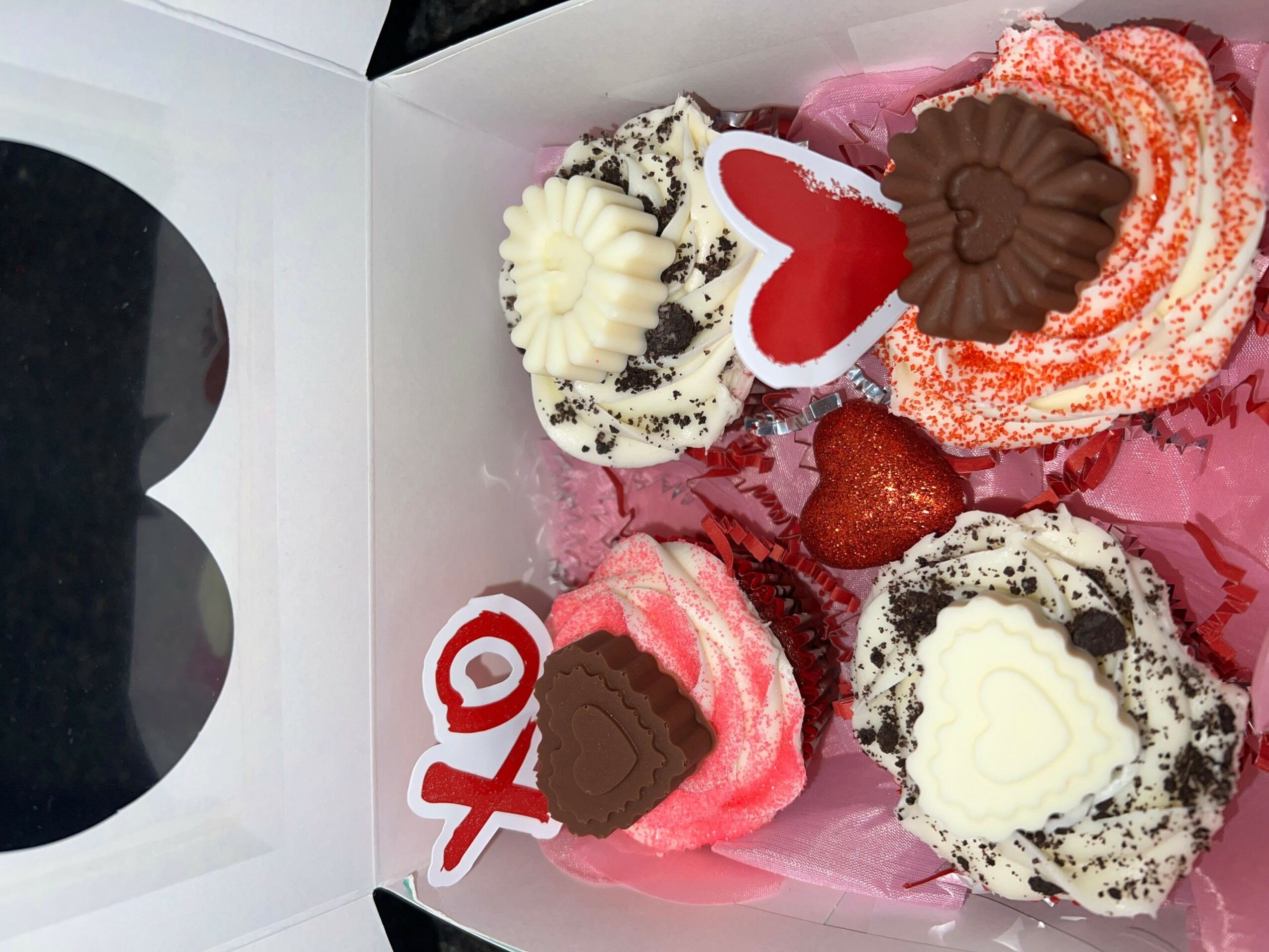Six decorated cupcakes with hearts and a white heart-shaped topper inside a box, some with pink and white frosting, some with chocolate, on pink paper